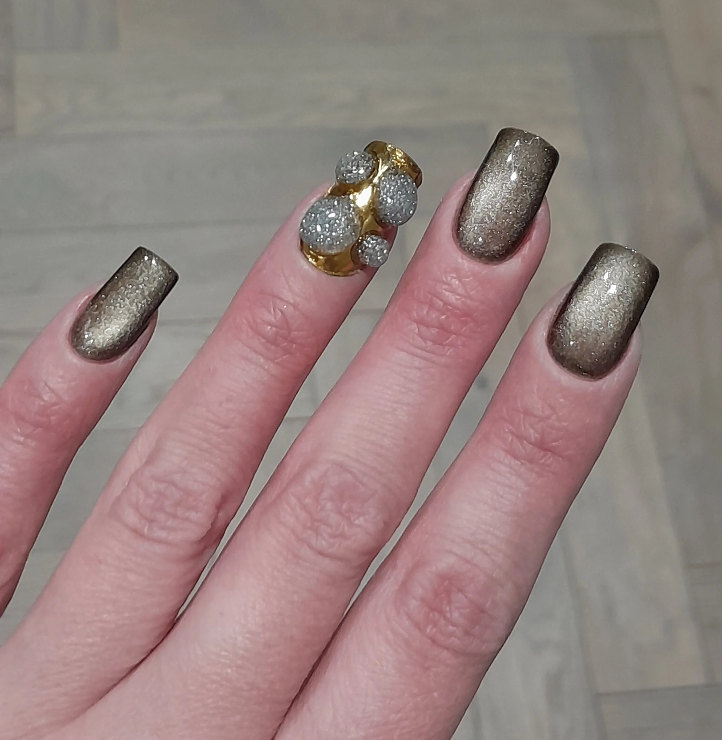 Close-up of a hand with painted nails in metallic gold and silver, wearing a gold ring with multiple diamond-studded spheres.