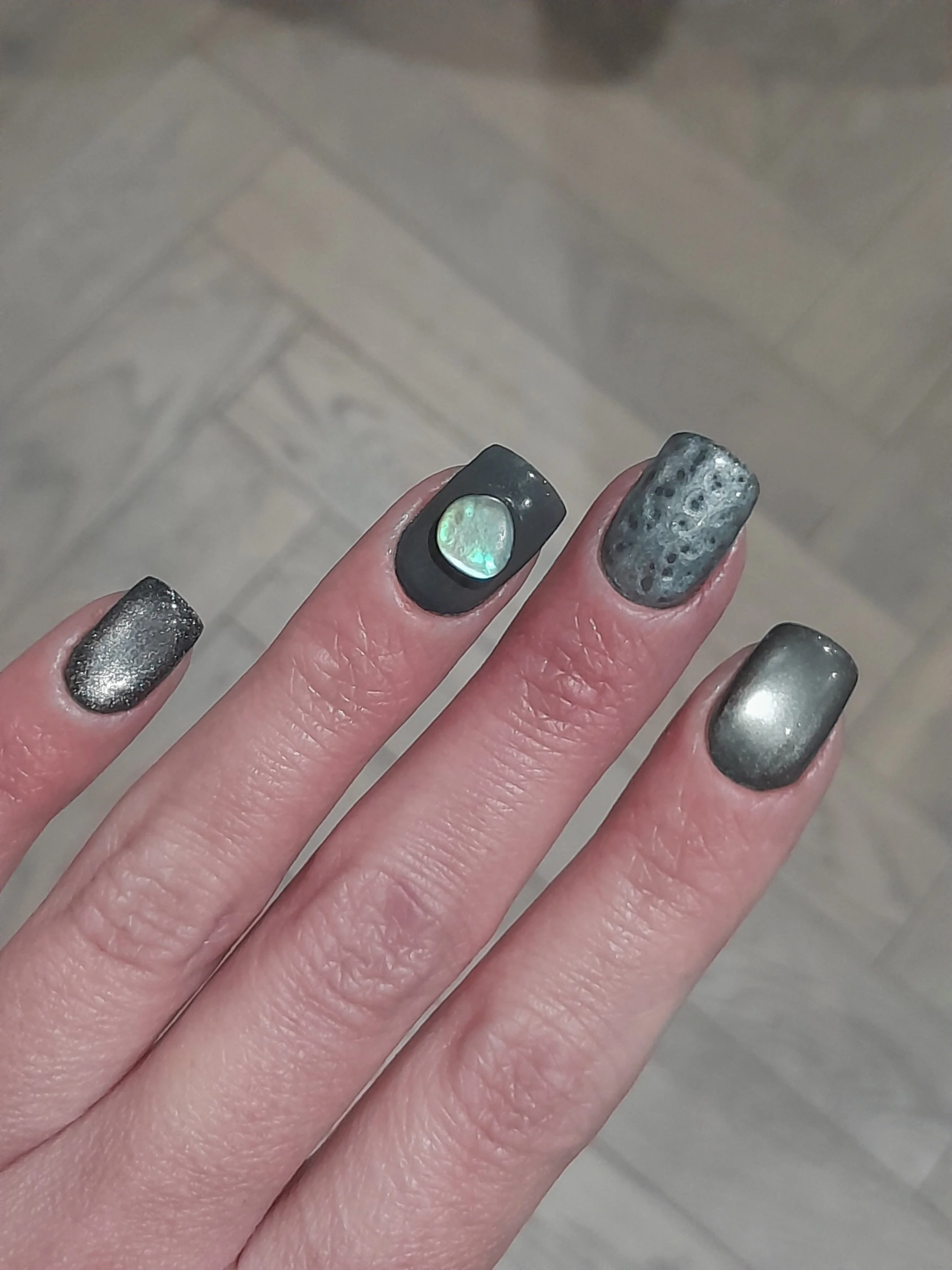 Close-up of a hand with five nails painted in metallic and black shades, featuring an abalone shell accent on the middle finger, against a background of wooden flooring.