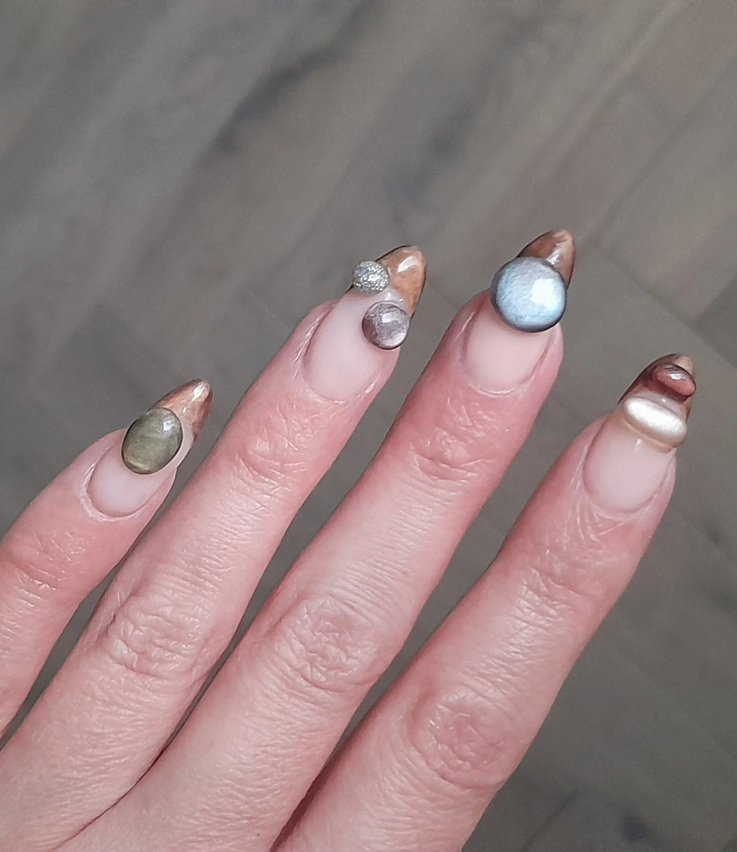 Close-up of a hand with nails painted in various colors and decorated with different embellishments, including a small diamond, metallic, and textured designs.