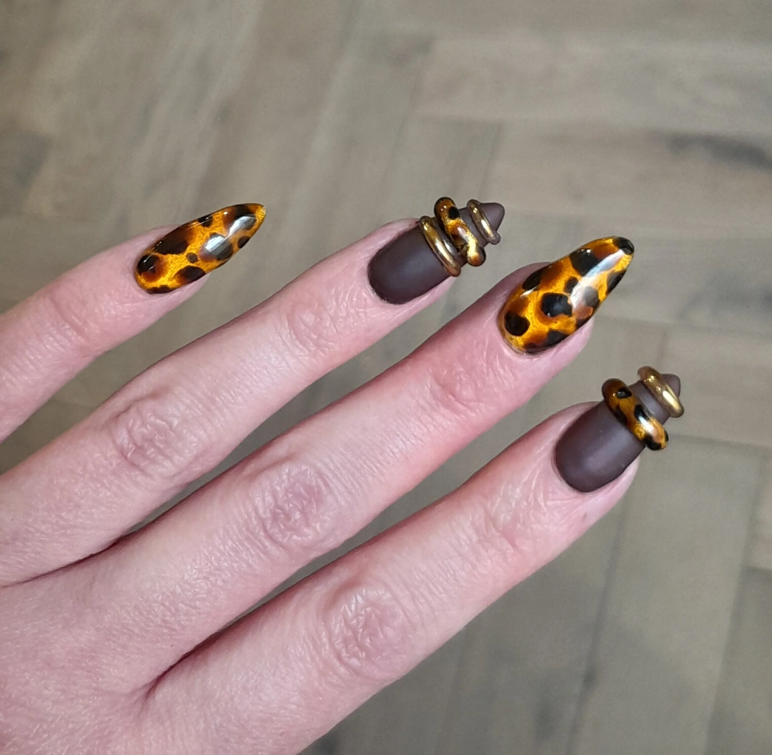Close-up of a hand with long painted nails featuring leopard print design and a matte brown base with gold and black accents.