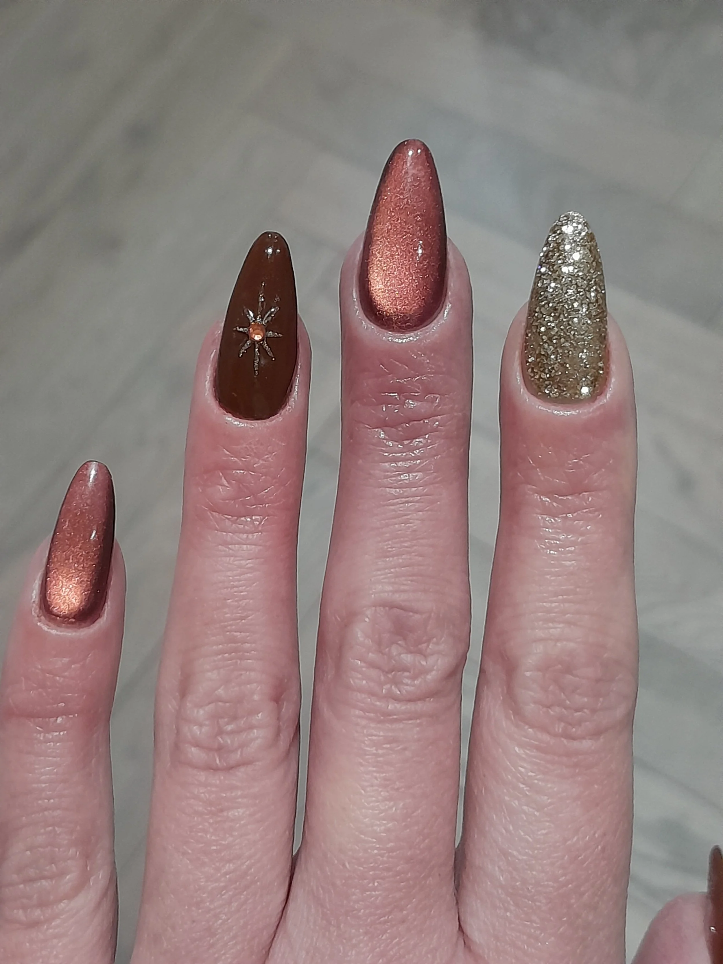 Close-up of a hand with five manicured fingernails, each painted with metallic or glittery nail polish in shades of bronze, gold, and copper.