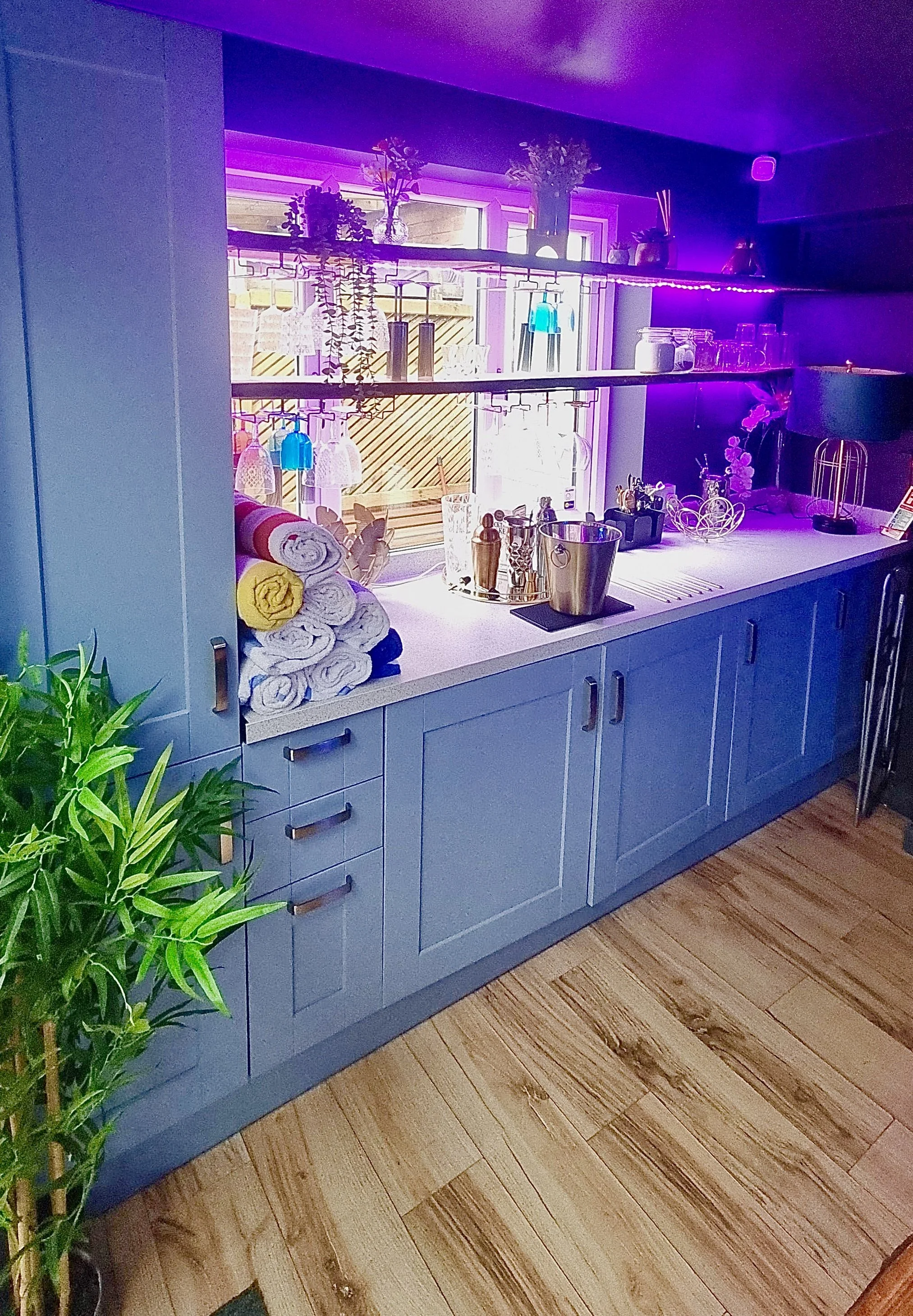A kitchen countertop with rolled towels, a potted plant on the left, and various bottles and decorative items under purple lighting on shelves above the window.