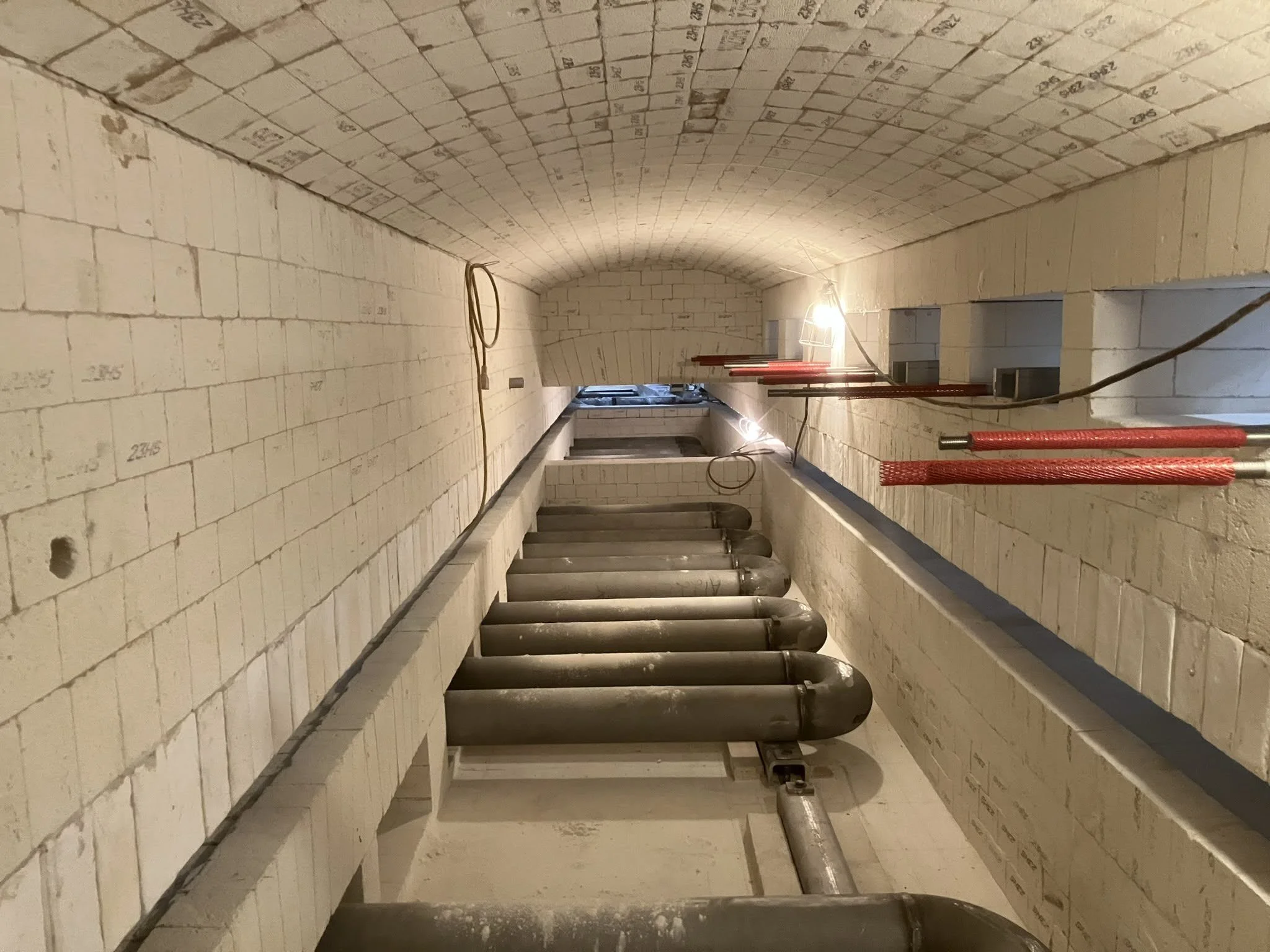 Underground tunnel with brick arched ceiling, concrete walls, and large pipes along the floor with some red safety barriers and electrical wiring.