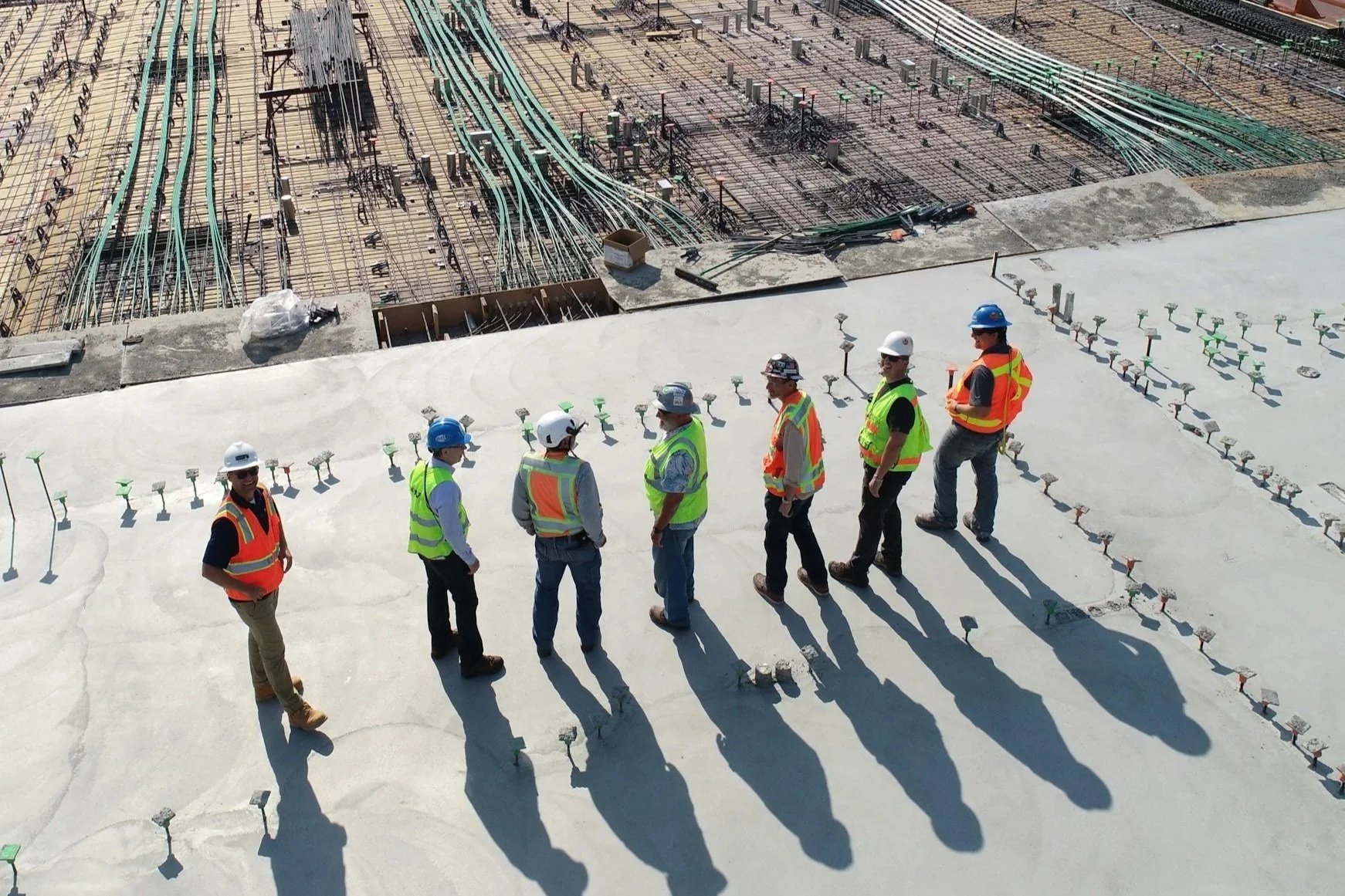 Group of construction workers in safety vests and helmets walking on a concrete roof with rebar and electrical wiring visible in the background.