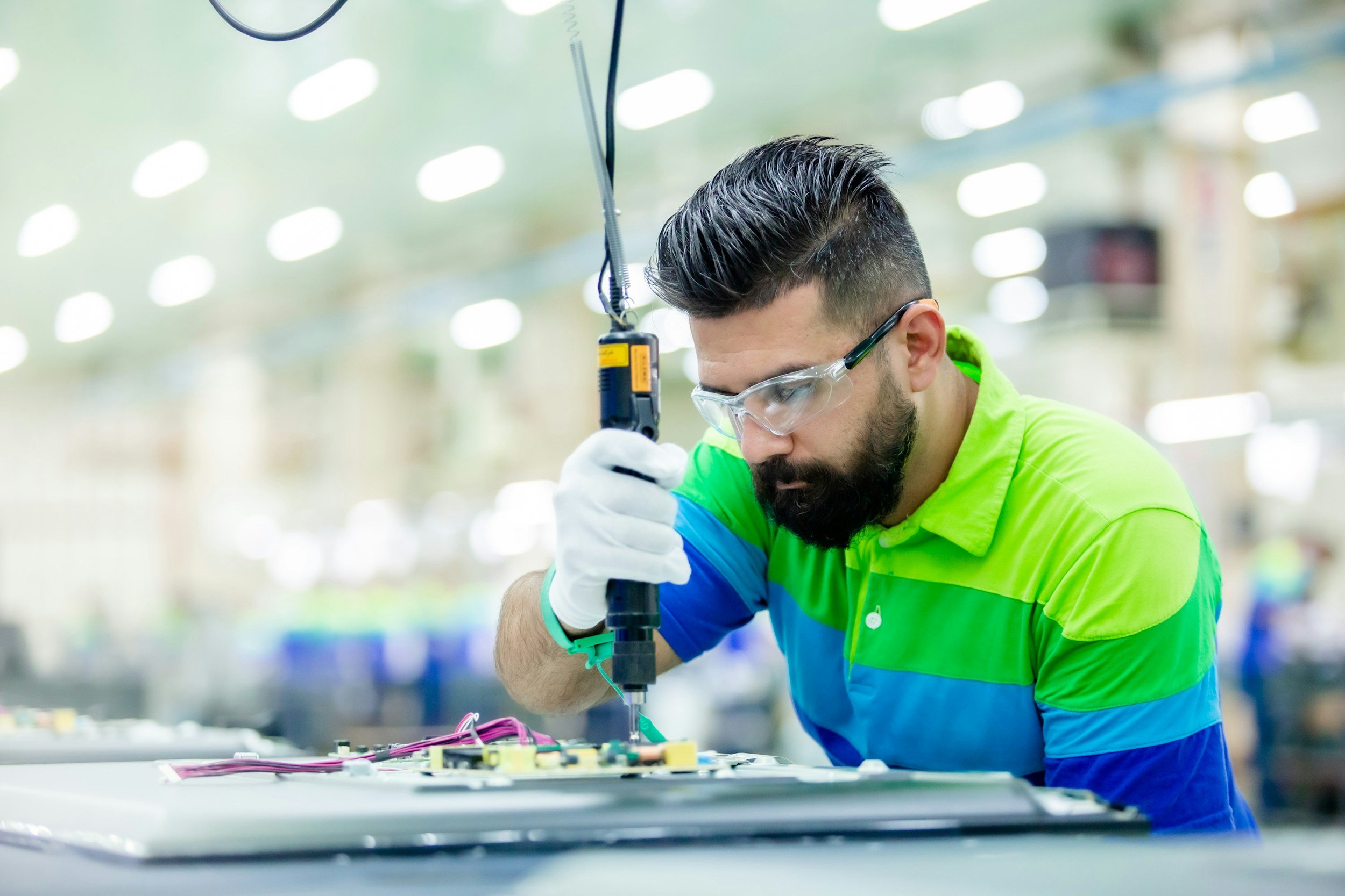 A man wearing safety glasses and gloves uses a screwdriver to work on electronic components in a workspace, with bright overhead lighting.