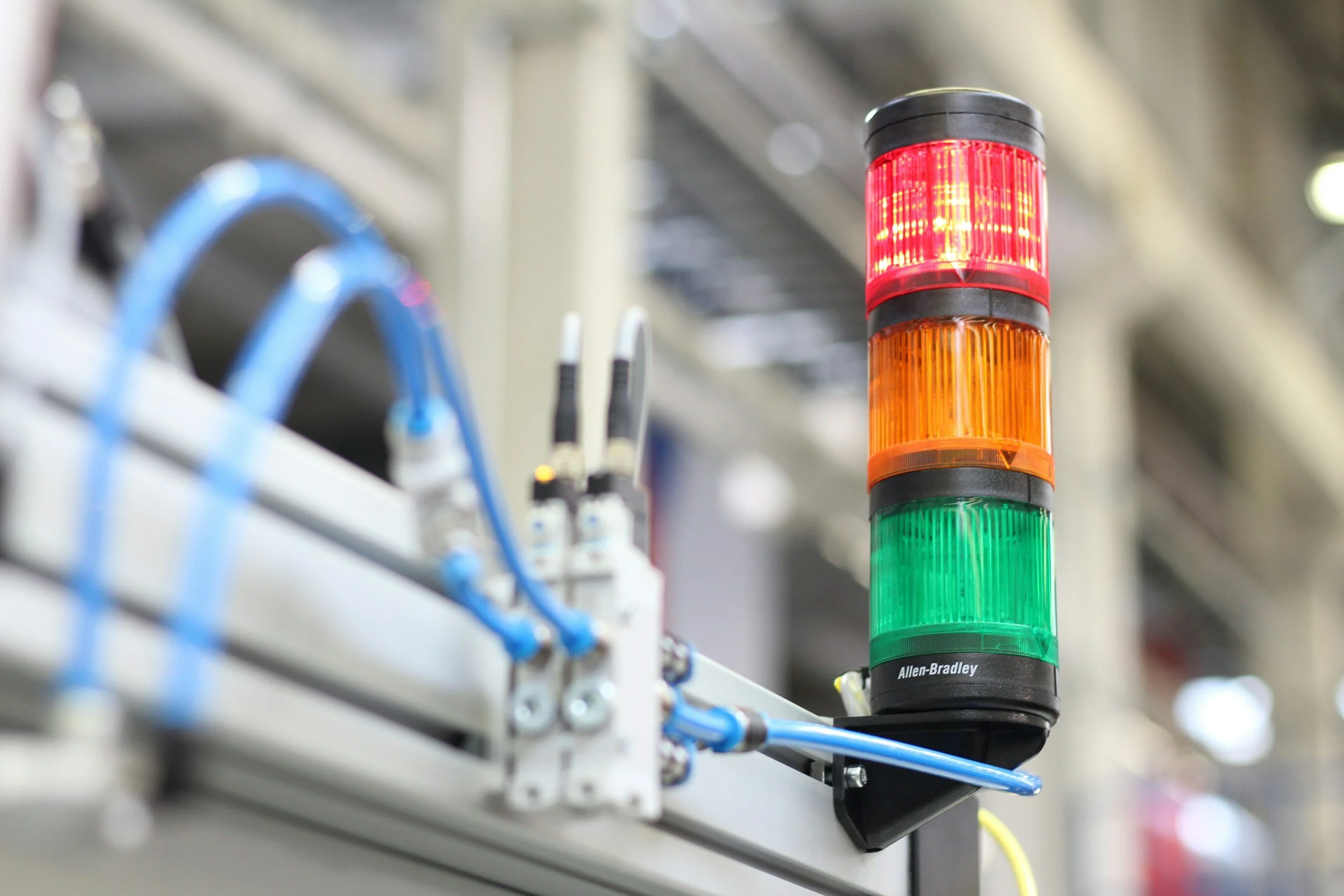 Industrial alarm light with red, orange, and green sections, mounted on a metal framework with blue and white wires in the background.