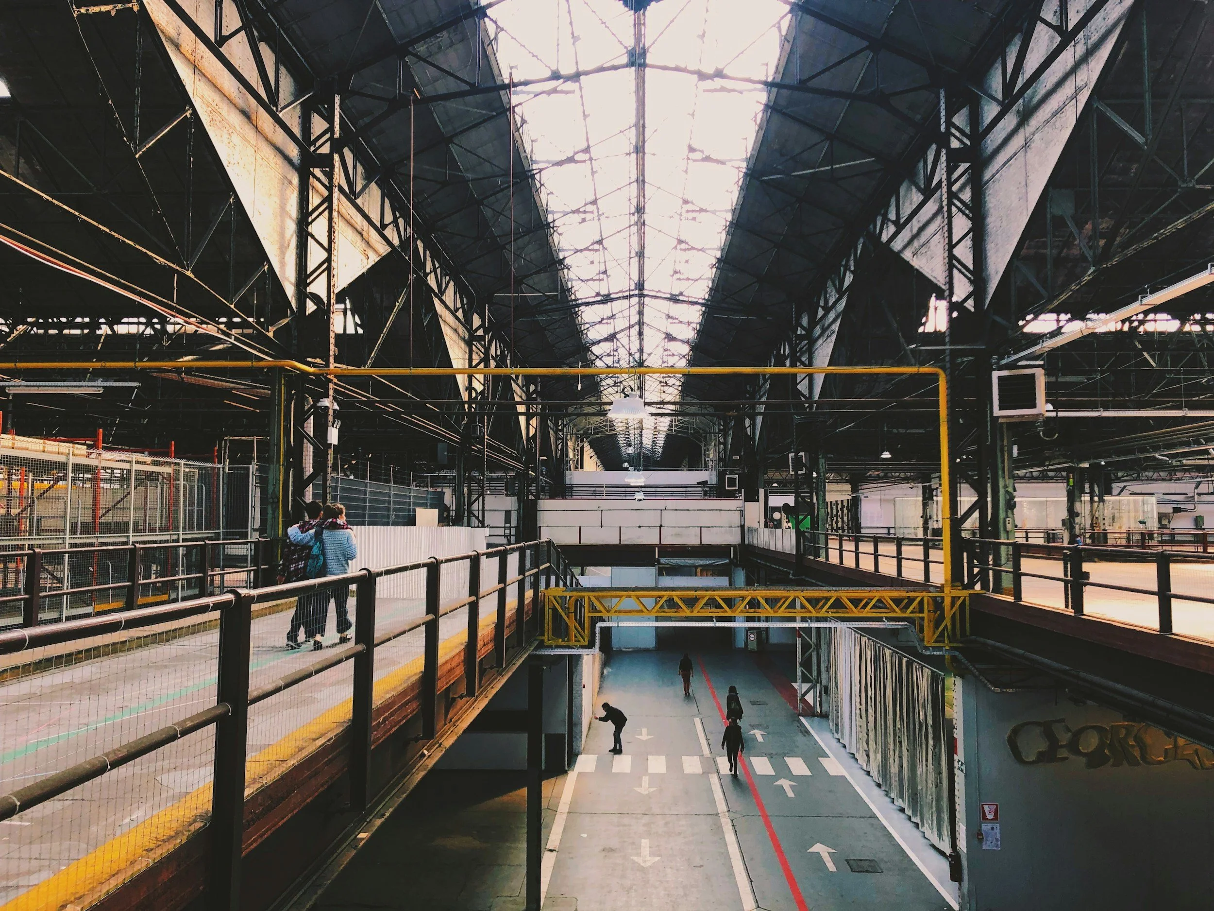 Inside an industrial building with high glass ceilings, steel frameworks, and multiple levels. A walkway with metal railings runs along the upper level. Several people are walking or standing, and the lower level has a pathway with arrows painted on the ground, leading to a closed gate and some graffiti on the wall.