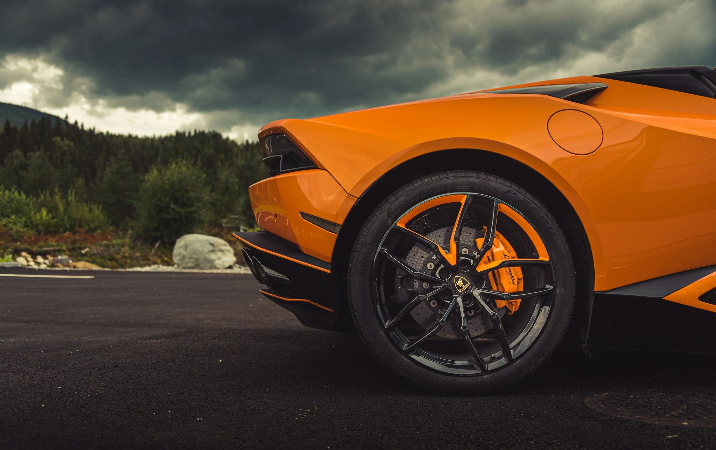 Close-up view of the front part of an orange sports car with black and orange rim, tire, brake caliper, and part of the front bumper, set against a cloudy sky and mountain landscape.