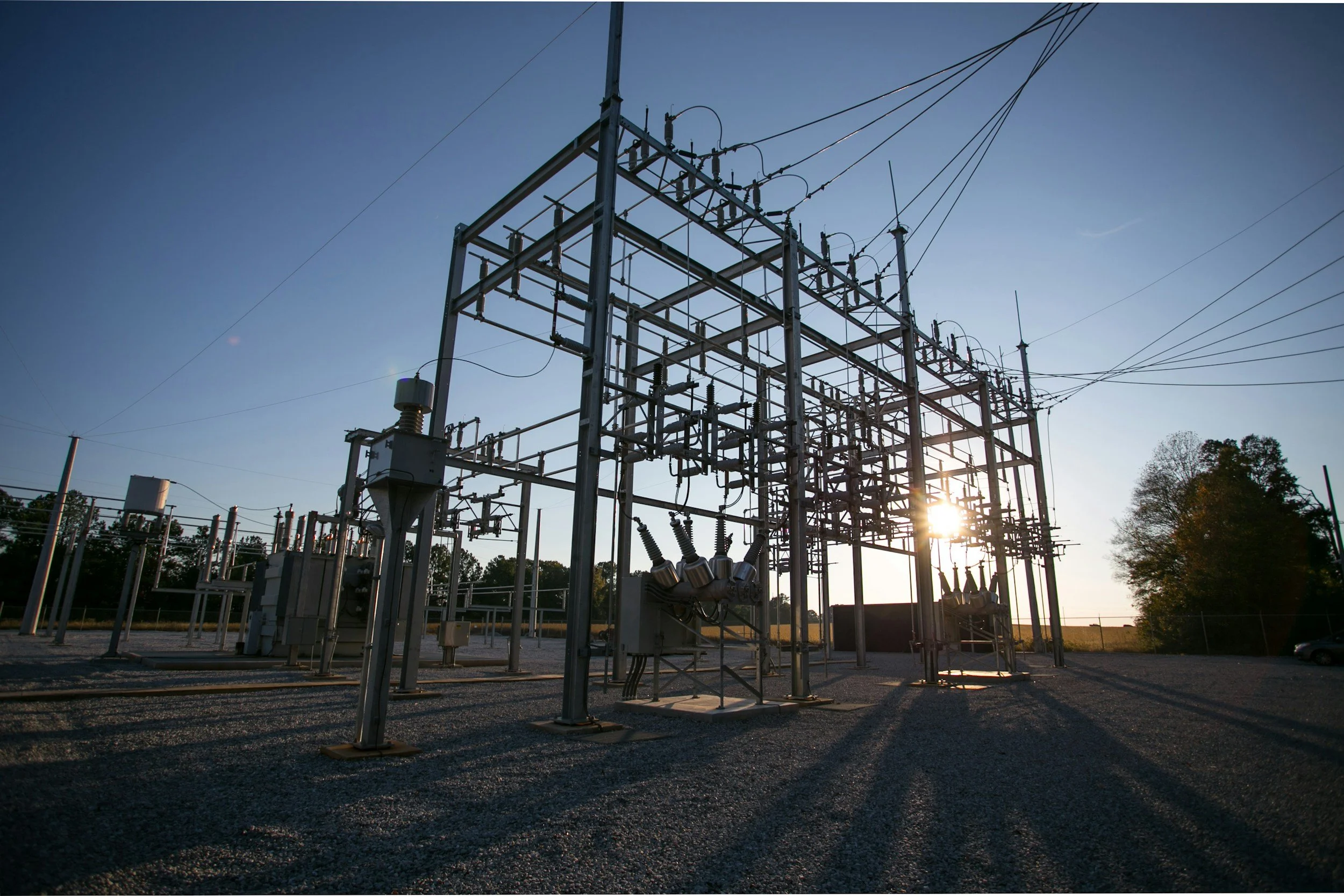 An outdoor electrical substation with metal structures, transformers, and power lines at sunset.