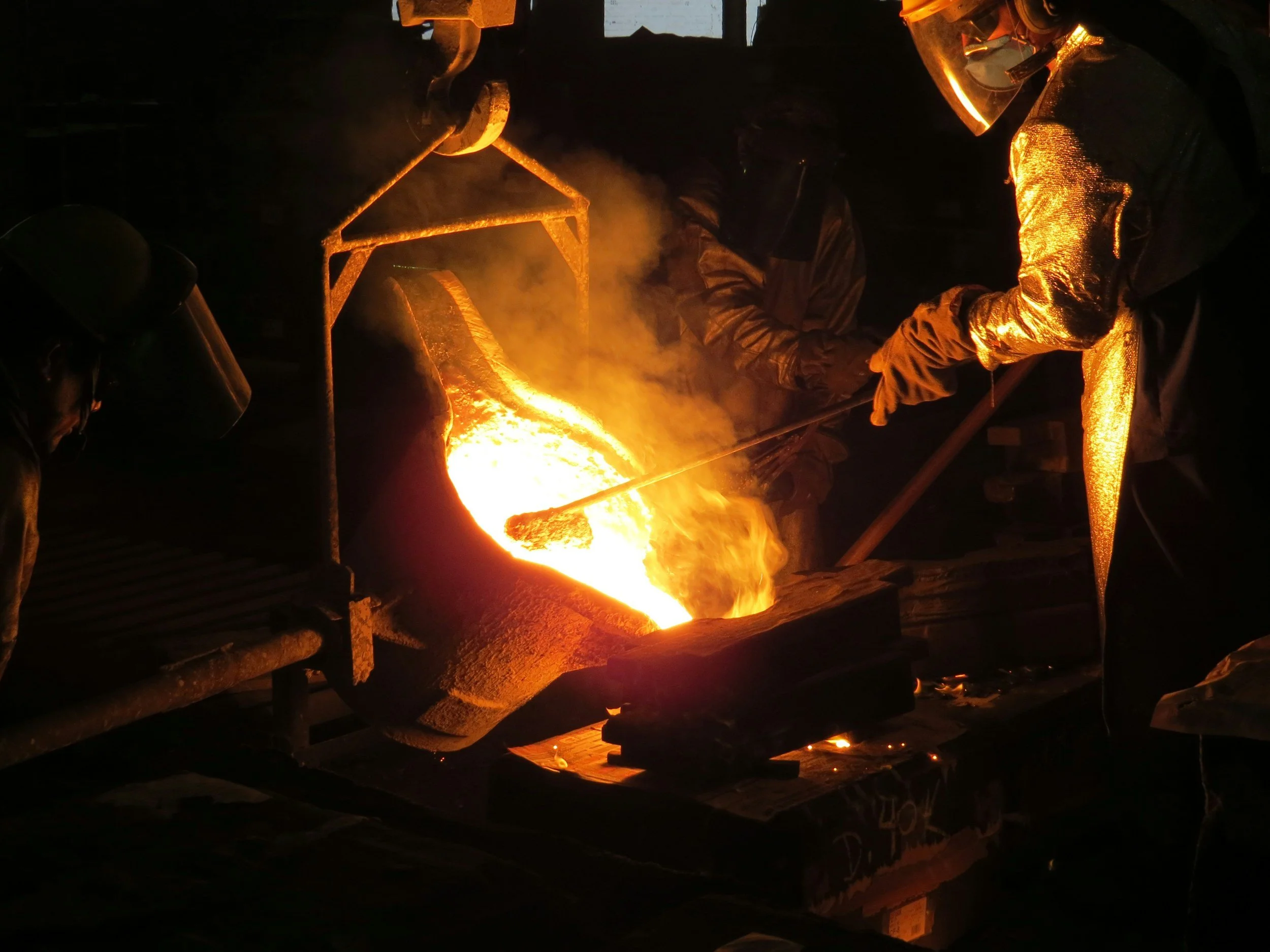 Three workers wearing protective gear are pouring molten metal from a large crucible into a mold during a foundry casting process, with bright orange flames and smoke visible.