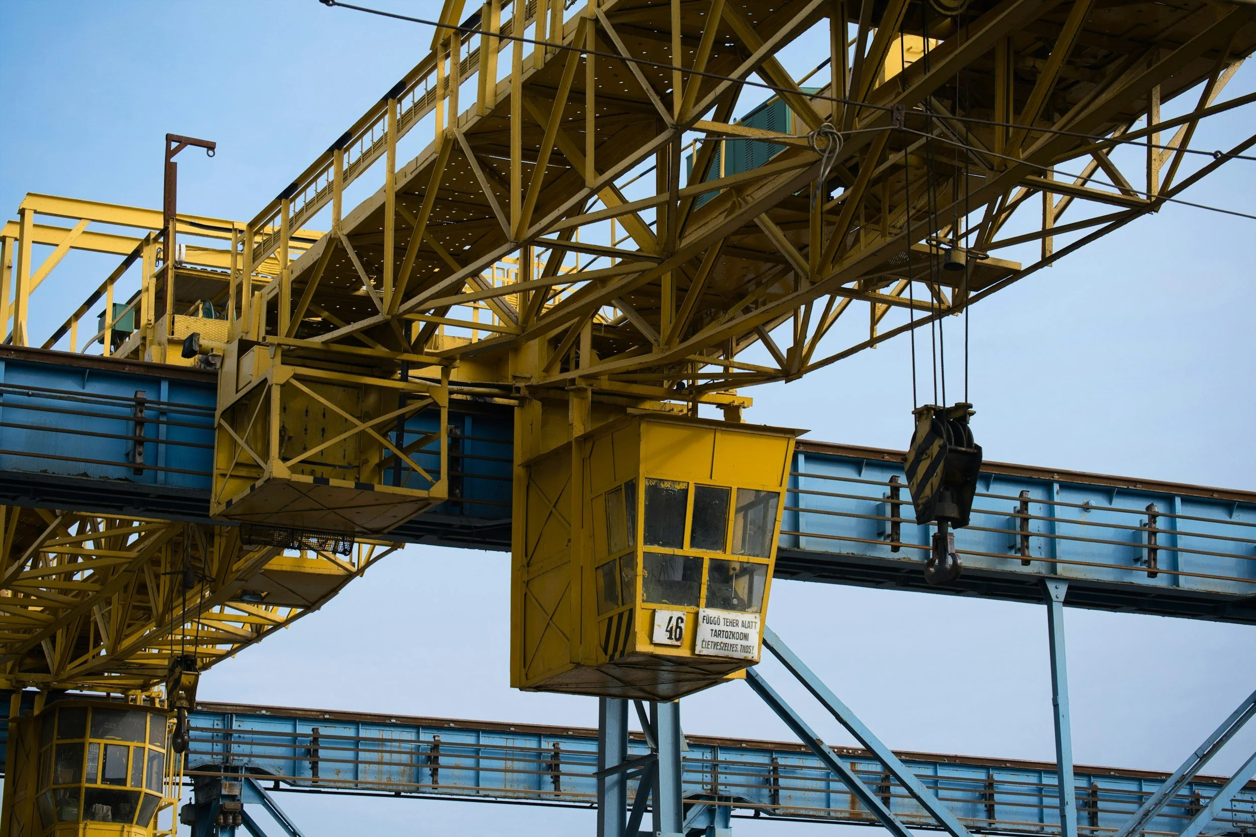 Close-up of a large industrial yellow and blue crane used for loading and unloading cargo with visible metal framework, pulley, and control cabin against a cloudy sky.