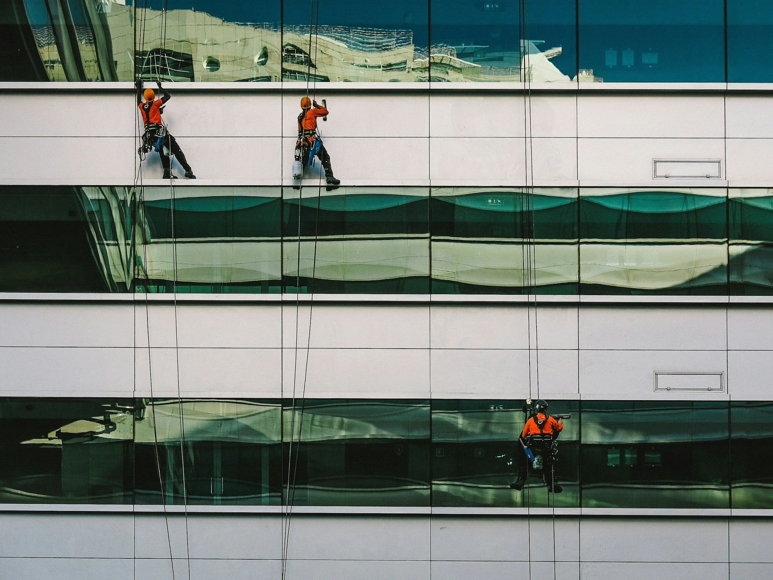 Three window washers cleaning the windows of a modern glass building with their safety harnesses and equipment.