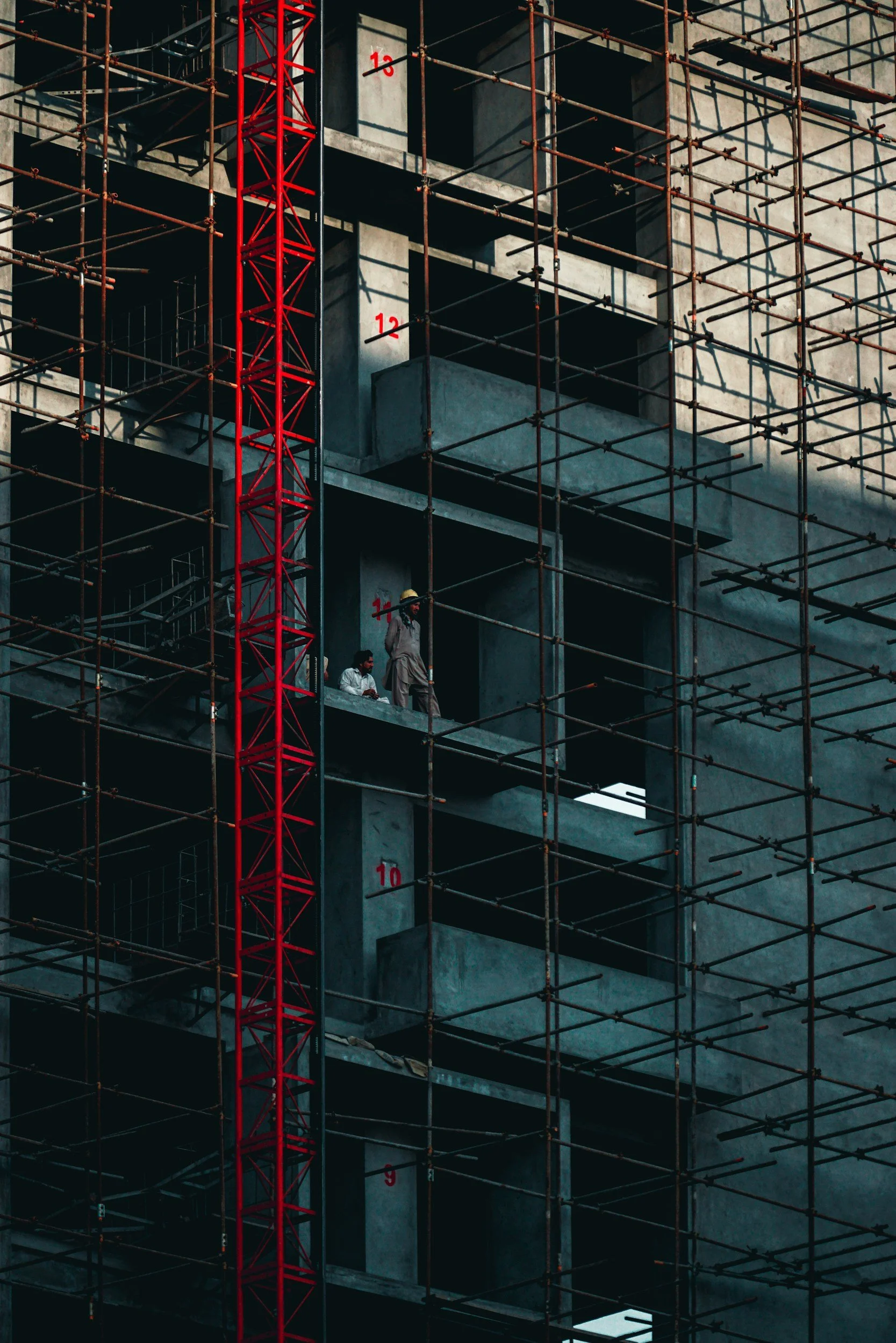 Construction site with scaffolding surrounding a multi-story building. Two workers are standing on a balcony, one wearing a yellow hard hat. The building has numbered markings from 9 to 13 on the concrete levels.