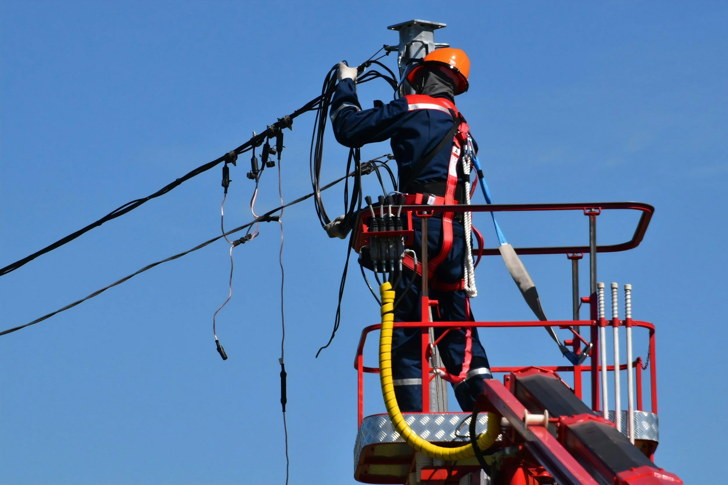 A worker in safety gear climbing a tall red lift bucket to repair or inspect electrical wires against a clear blue sky.