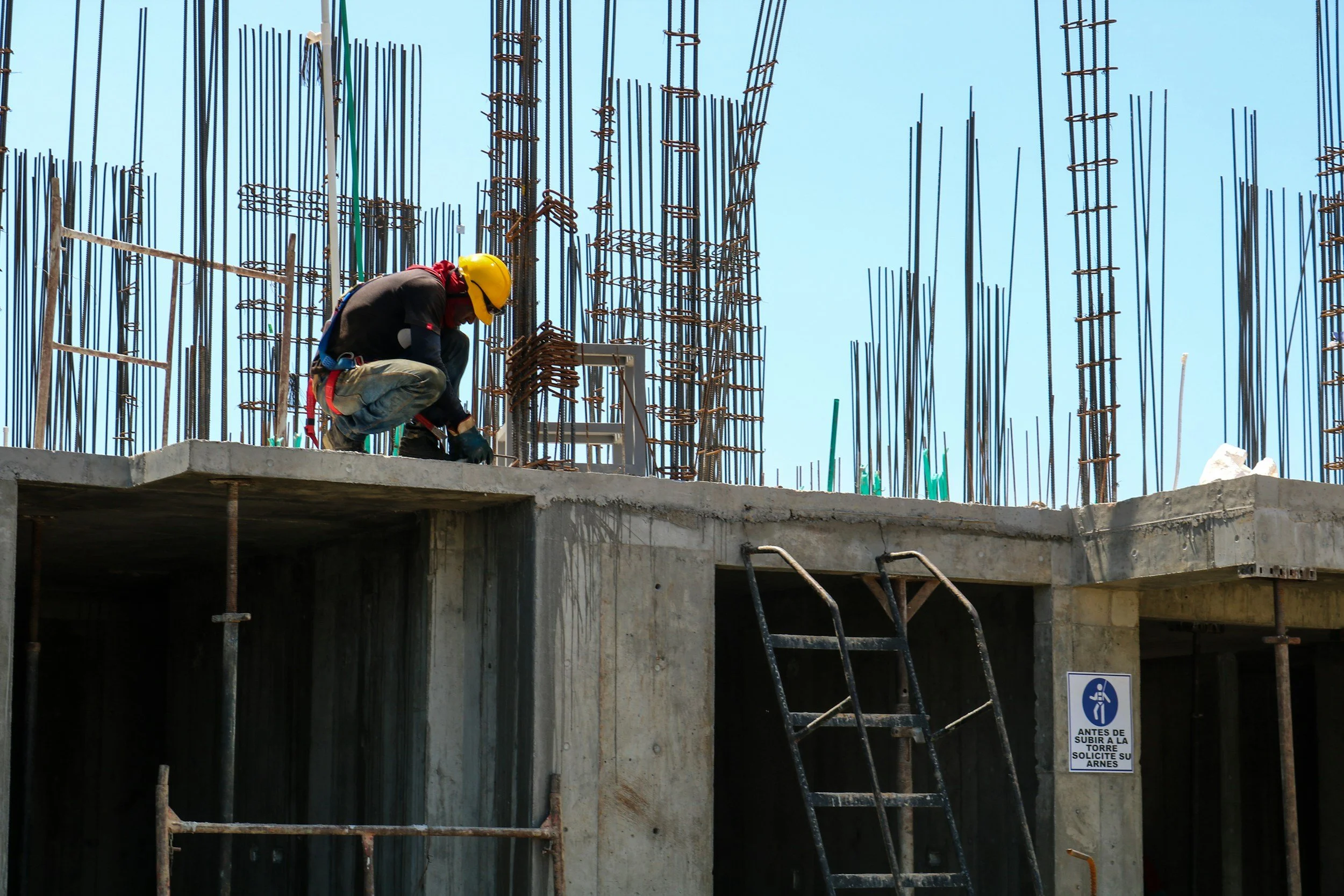 A construction worker wearing a yellow safety helmet is crouched on a concrete structure, working with rebar at a building site with a steel ladder leaning against it and a warning sign in Spanish.