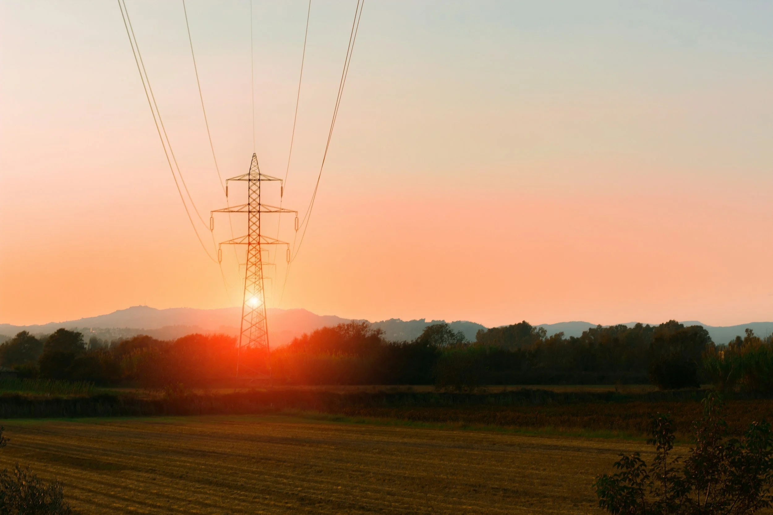 A sunset with an orange and pink sky, a power line tower, and rolling hills with trees in the background.