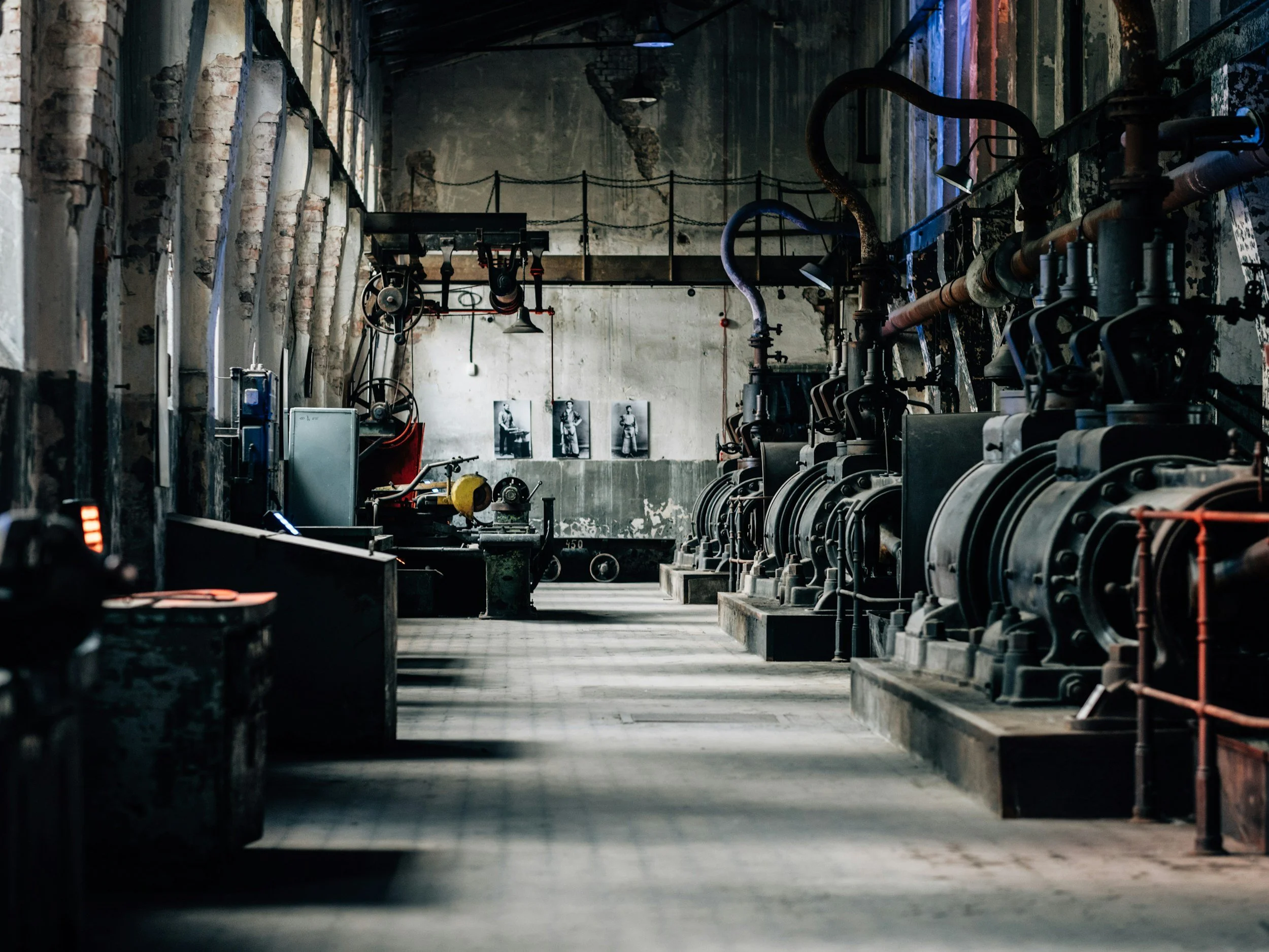 An industrial factory interior with large machinery and equipment, exposed pipes, and black and white photos on the back wall.