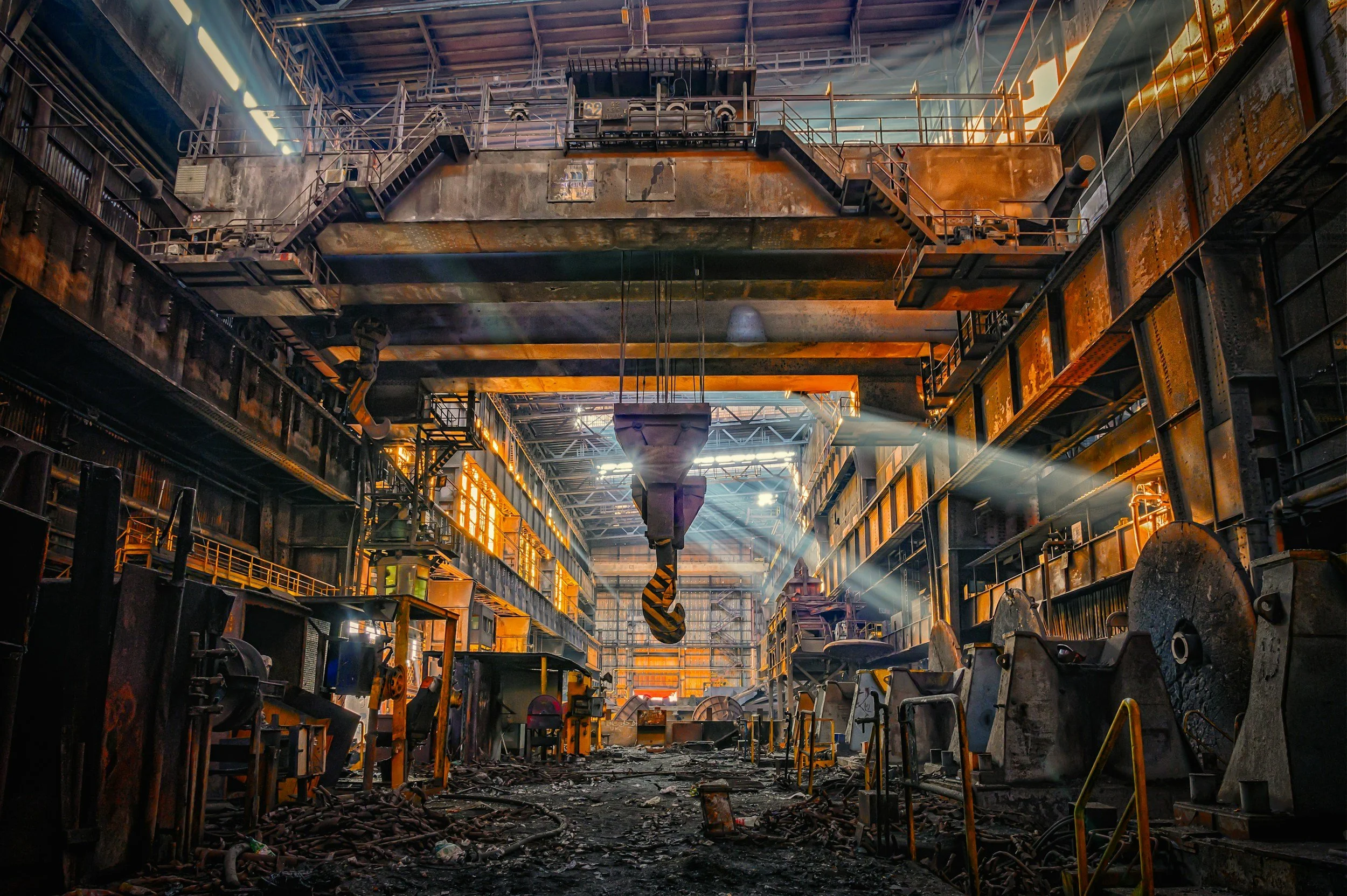 Interior of an abandoned, rusted industrial factory with large machinery, overhead cranes, and debris on the floor, illuminated by sunlight streaming through windows.