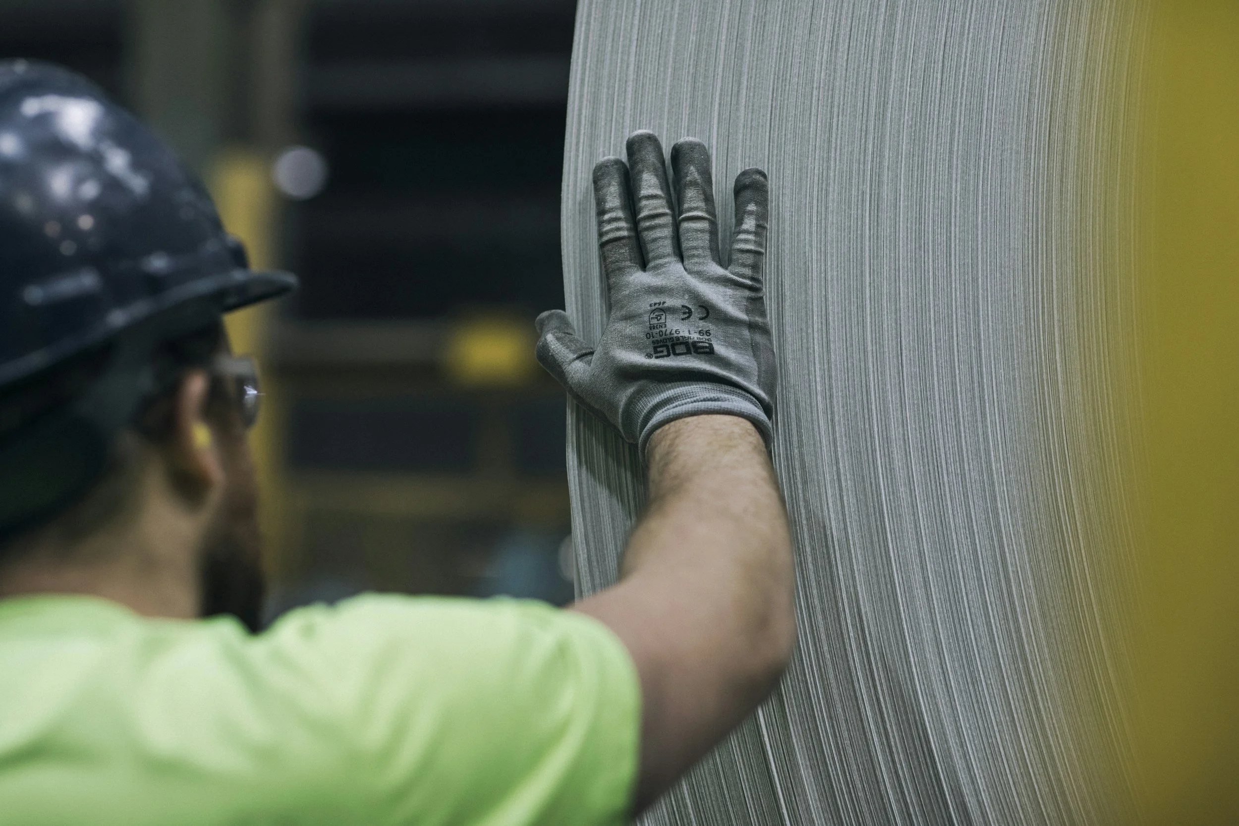 A construction worker wearing a black safety helmet and gray work gloves touches a large, textured metal sheet with his left hand.