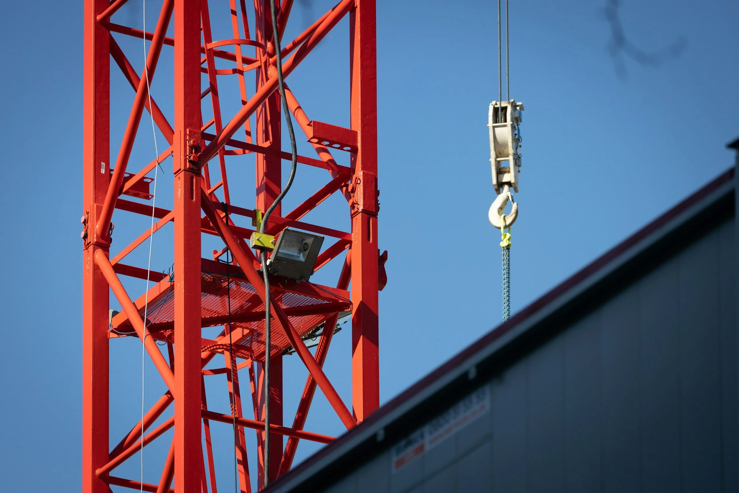 Close-up of a red construction tower crane with a hook hanging from a cable against a clear blue sky.