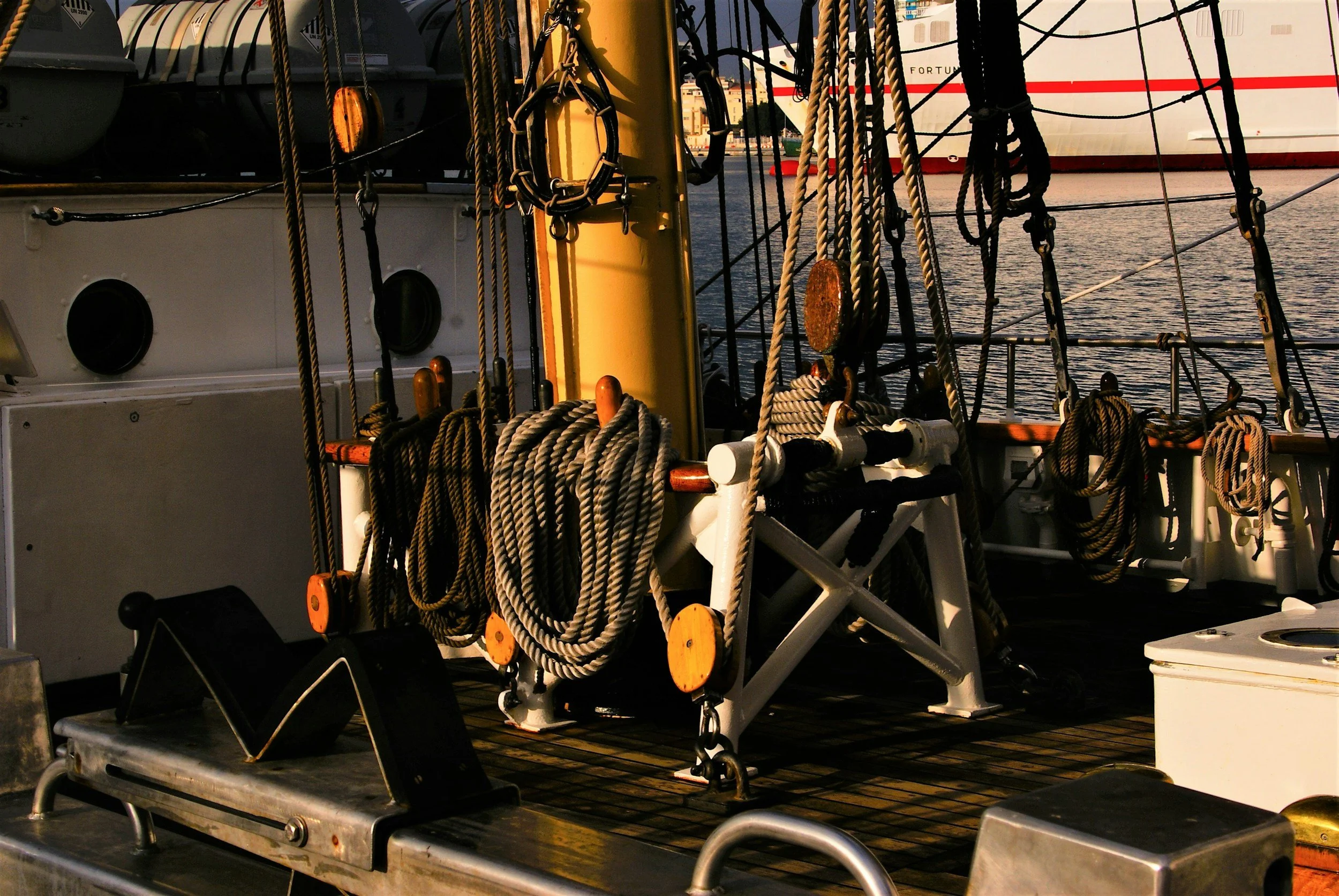Close-up of a ship's deck showing coiled ropes, pulleys, and rigging, with water and another ship in the background at sunset.