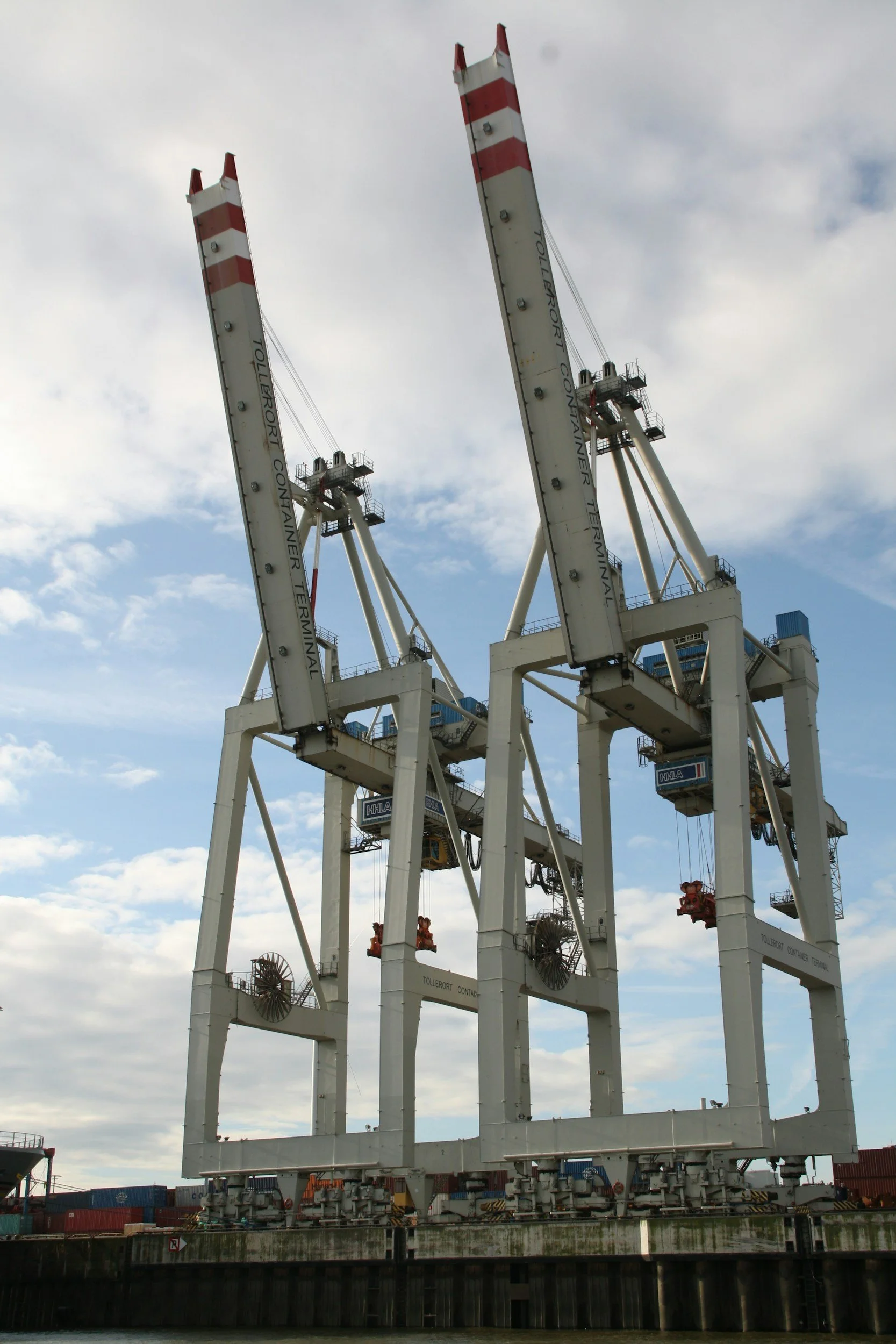 A large industrial container crane at a port, with two tall booms extending upwards against a partly cloudy sky.