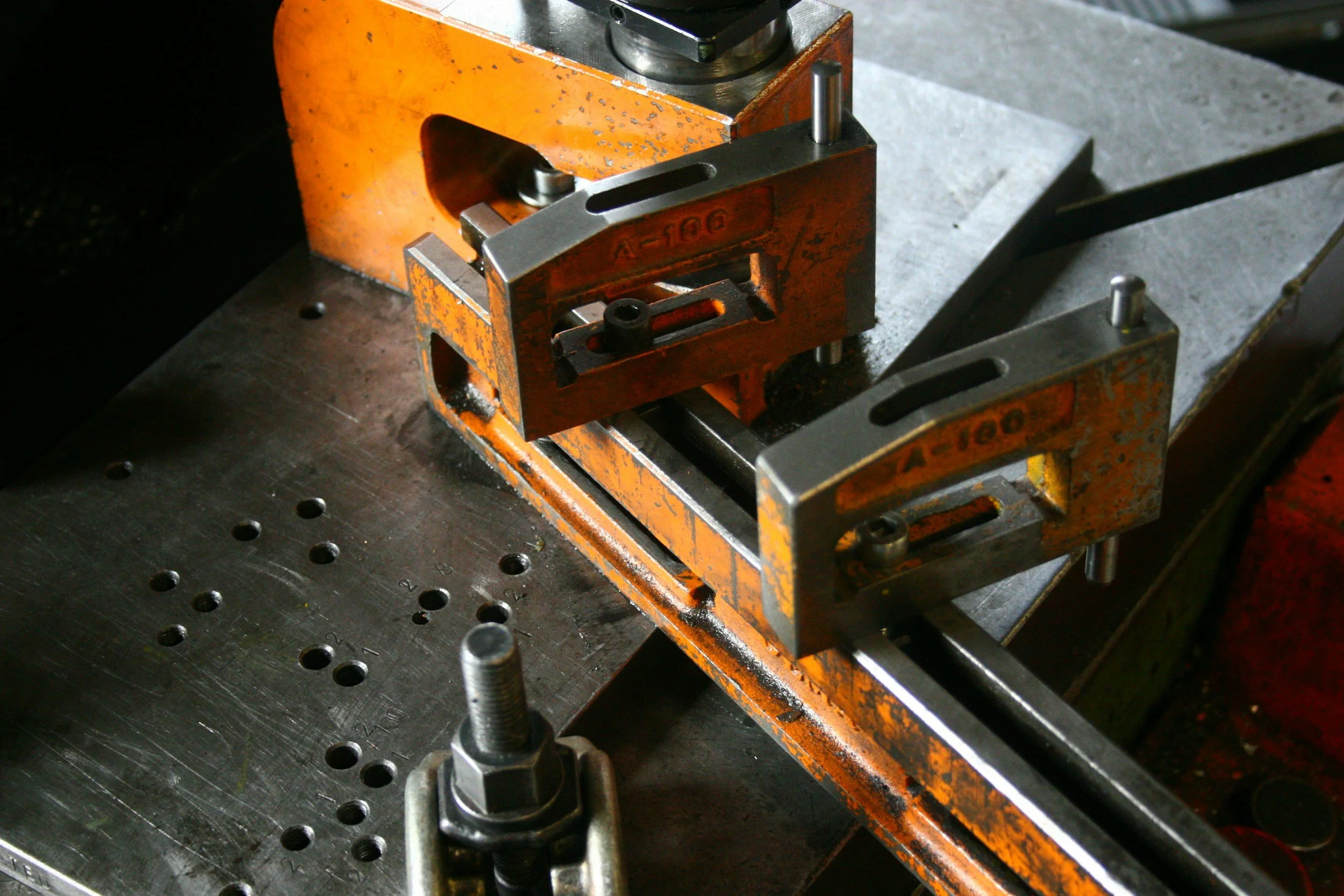 Two orange metal clamps securing metal pieces on a workbench with a drill and a metal plate with holes.
