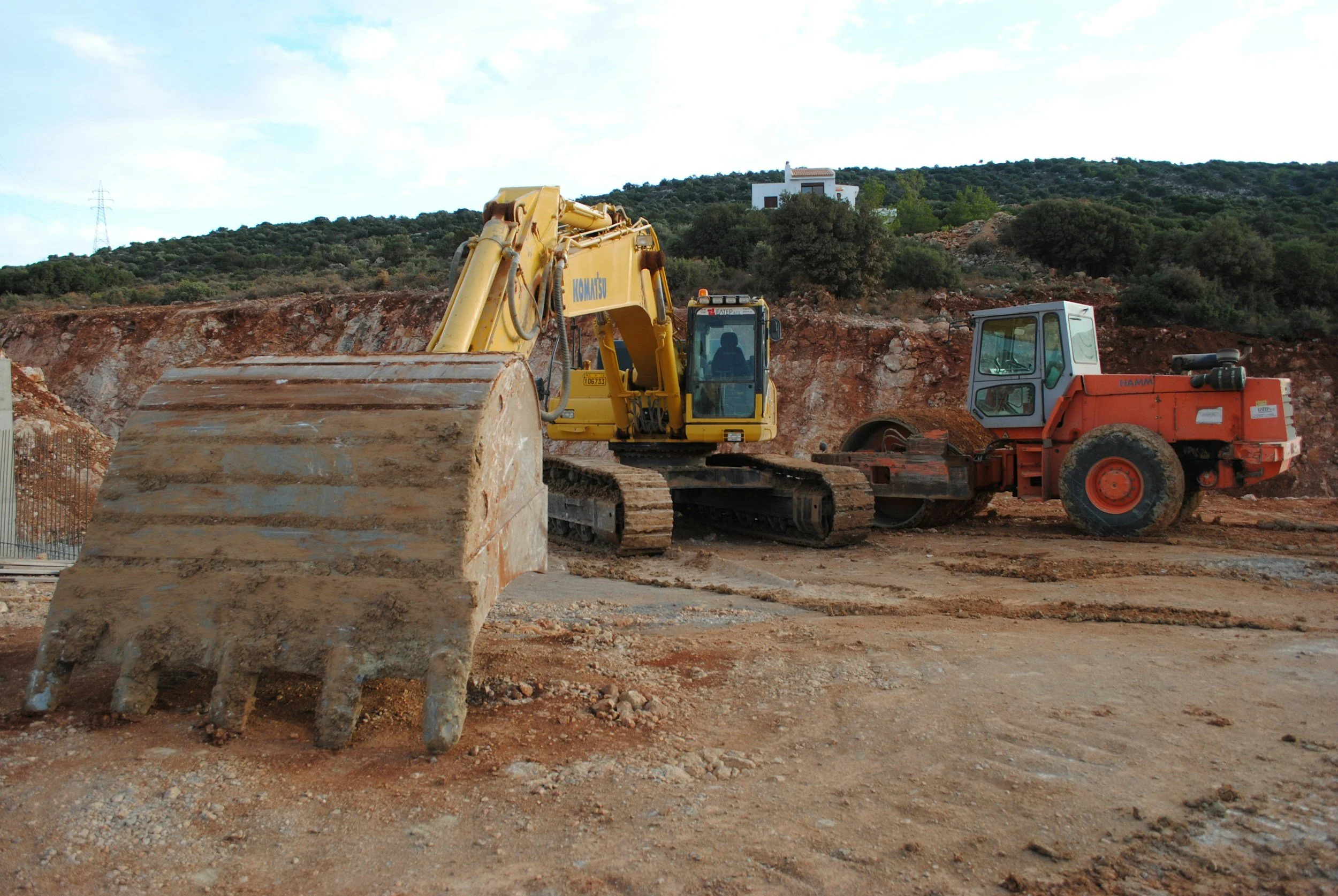 A yellow excavator and a red steamroller at a construction site on a dirt surface with a hillside and trees in the background.