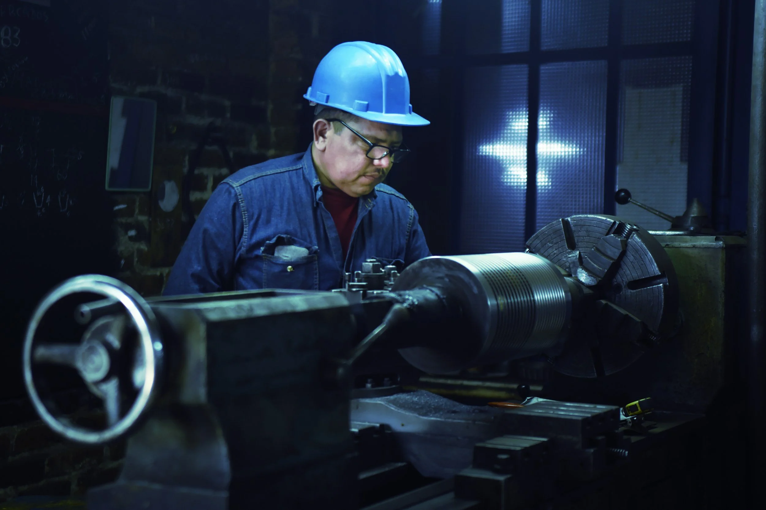 A worker wearing a blue hard hat and safety glasses operating a large lathe machine in a workshop.
