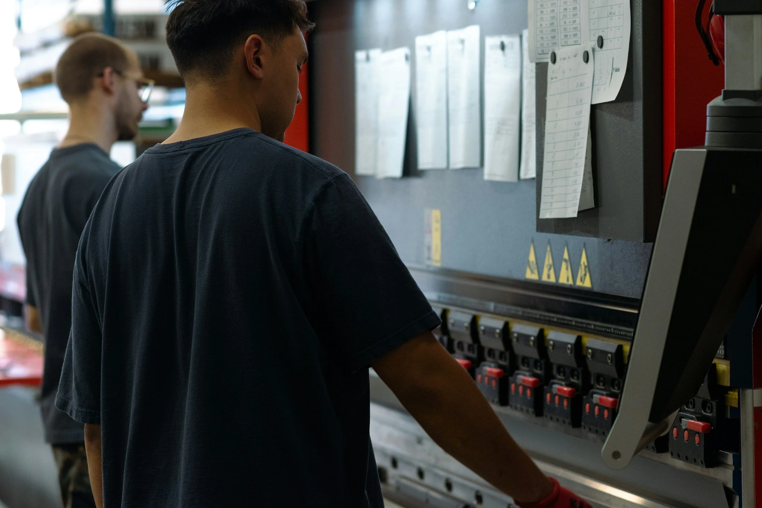 Two workers in a warehouse operating a large electrical panel with switches and papers attached to a bulletin board.
