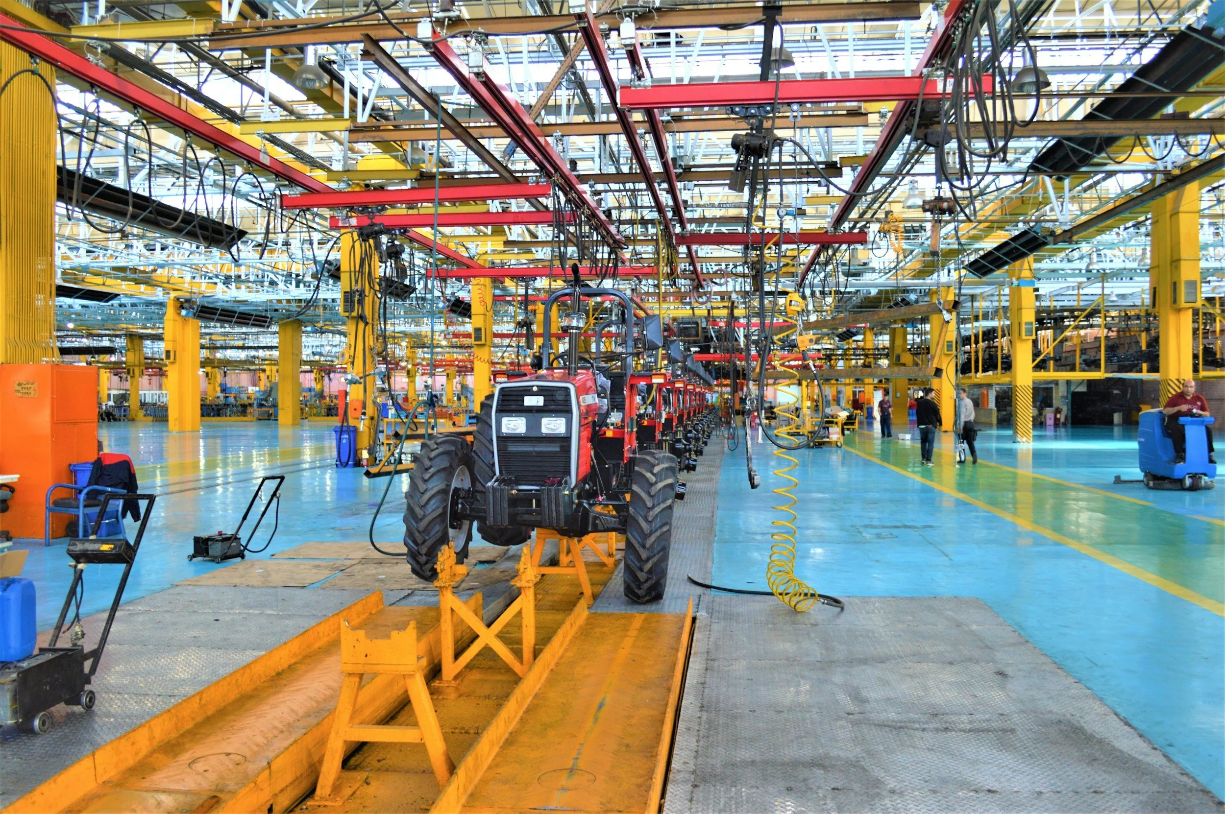 Inside a large industrial factory or assembly plant, with yellow support beams and various machinery, including a red tractor on a maintenance track, and workers in the background.