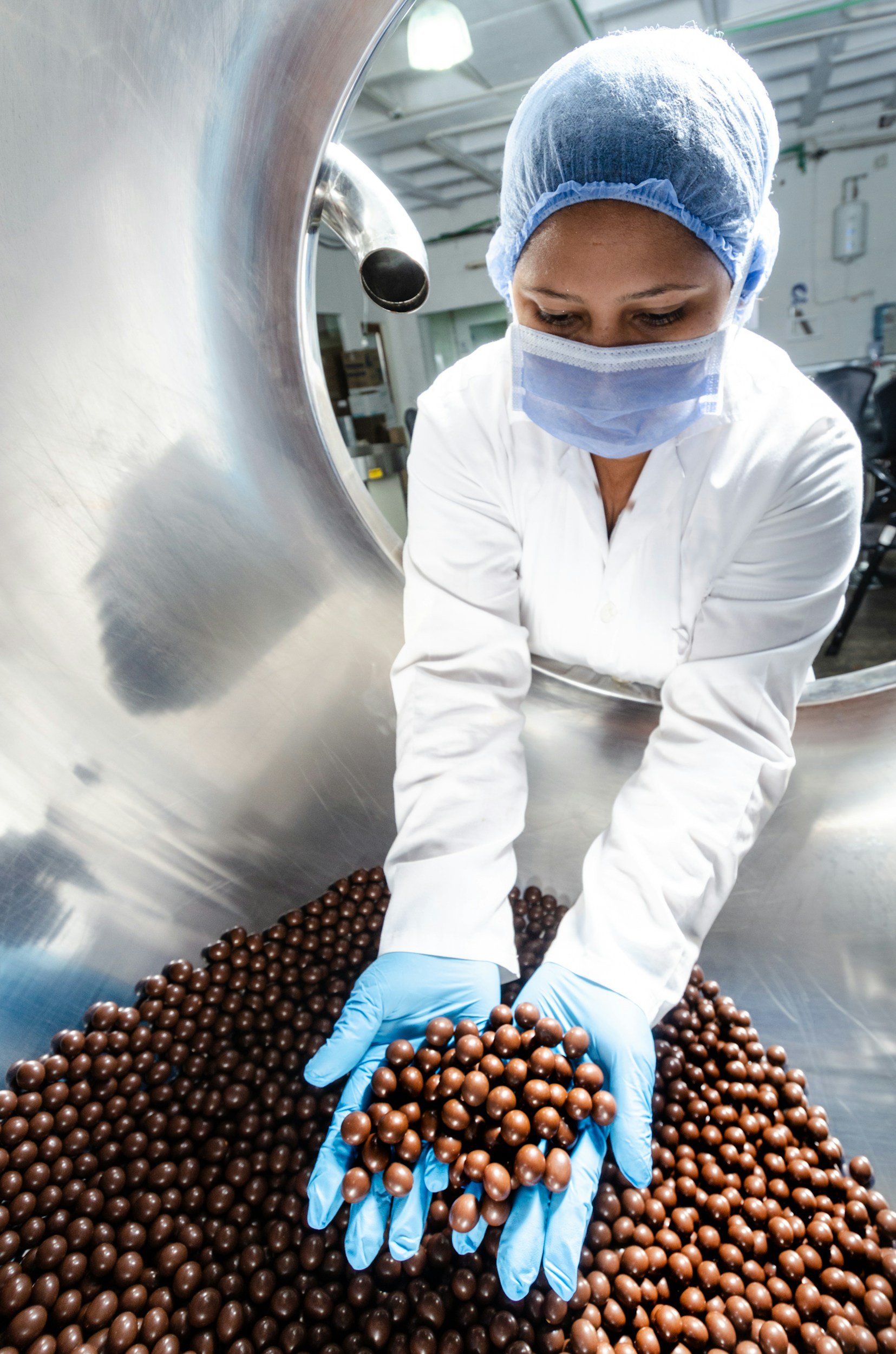 A person wearing a white coat, blue gloves, a face mask, and a hairnet handling roasted chocolate nibs or coffee beans in a large metal container.