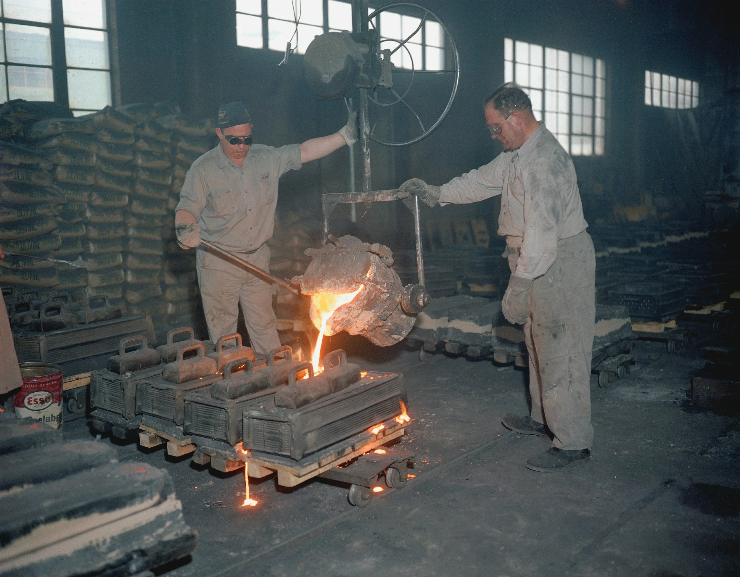 Two workers in a foundry pouring molten metal from a large crucible into molds on a mobile cart, with stacks of molds and industrial equipment in the background.