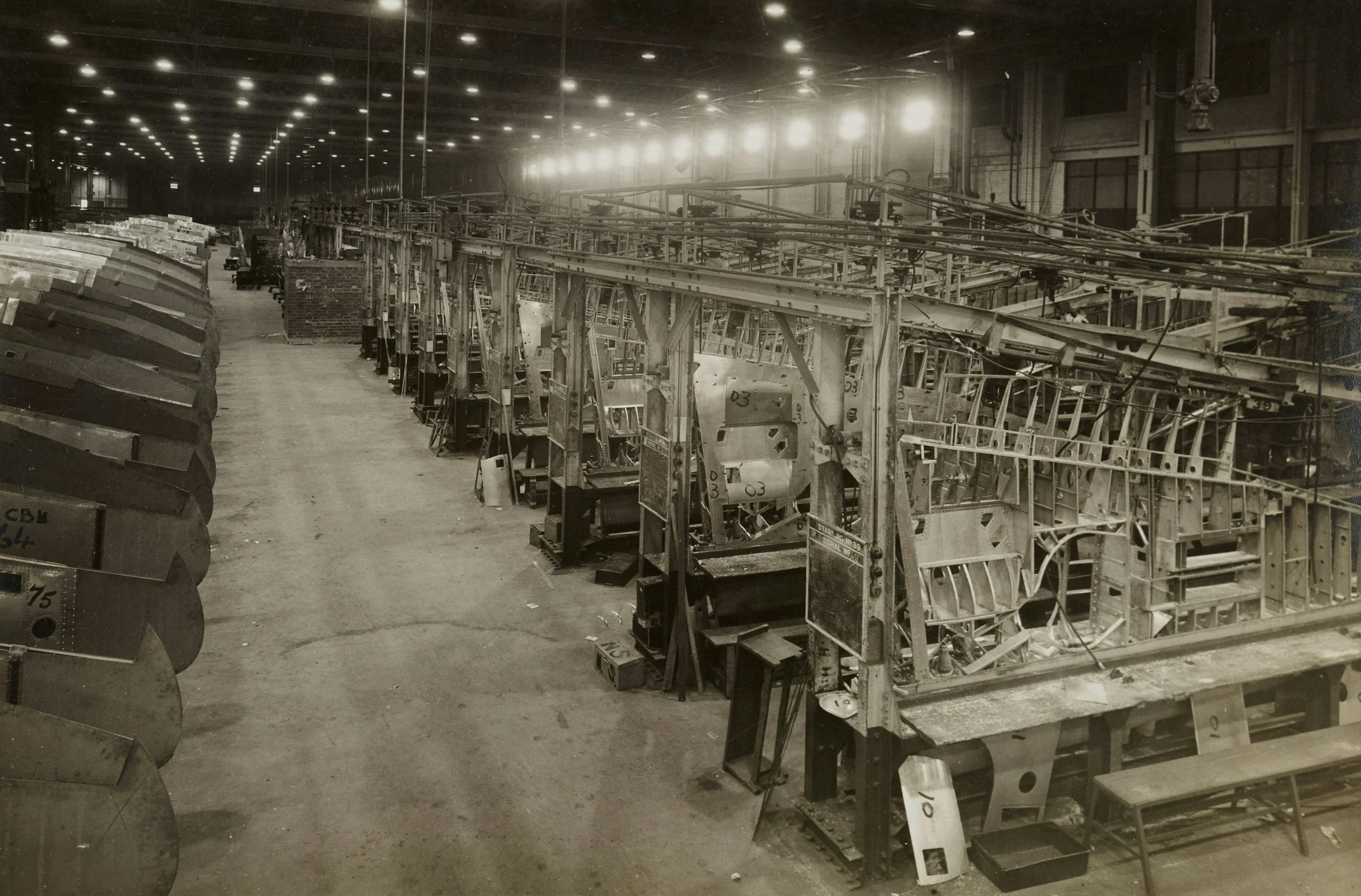 A large, empty industrial factory or manufacturing plant with rows of disassembled aircraft fuselages and large machinery, illuminated by overhead lights.