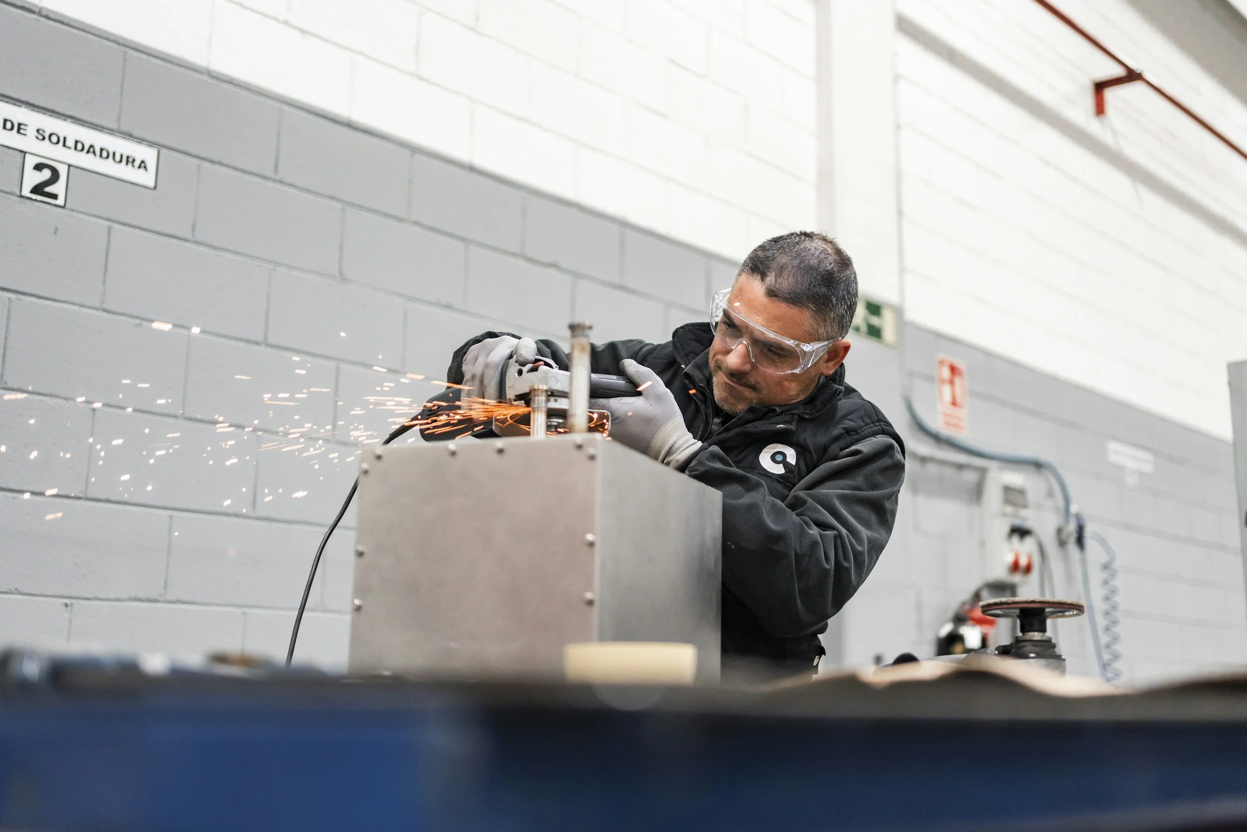 A man wearing safety goggles and gloves is using a tool to cut or grind a metal piece, creating sparks in an industrial workshop.