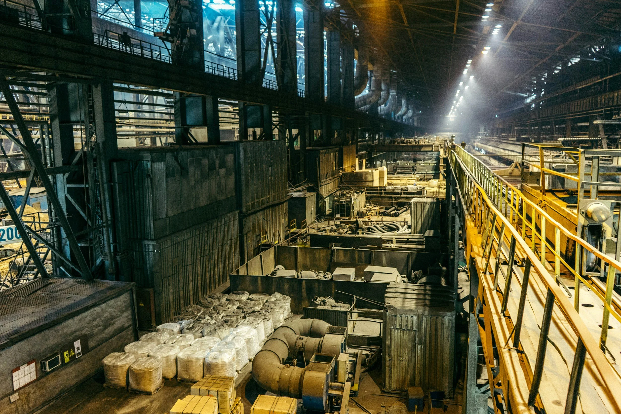 Interior of an industrial factory or warehouse with metal structures, machinery, and yellow safety railing along a walkway.
