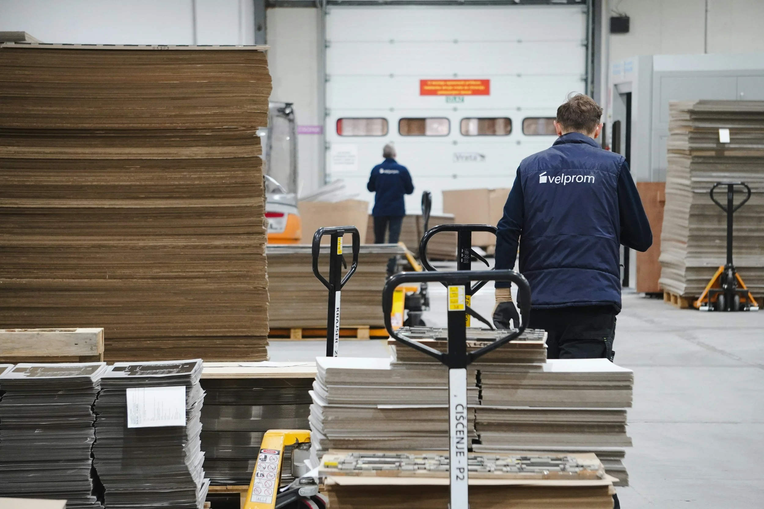 Workers in a warehouse handling large stacks of cardboard and paper, with forklifts and pallets visible, and a white garage door in the background.