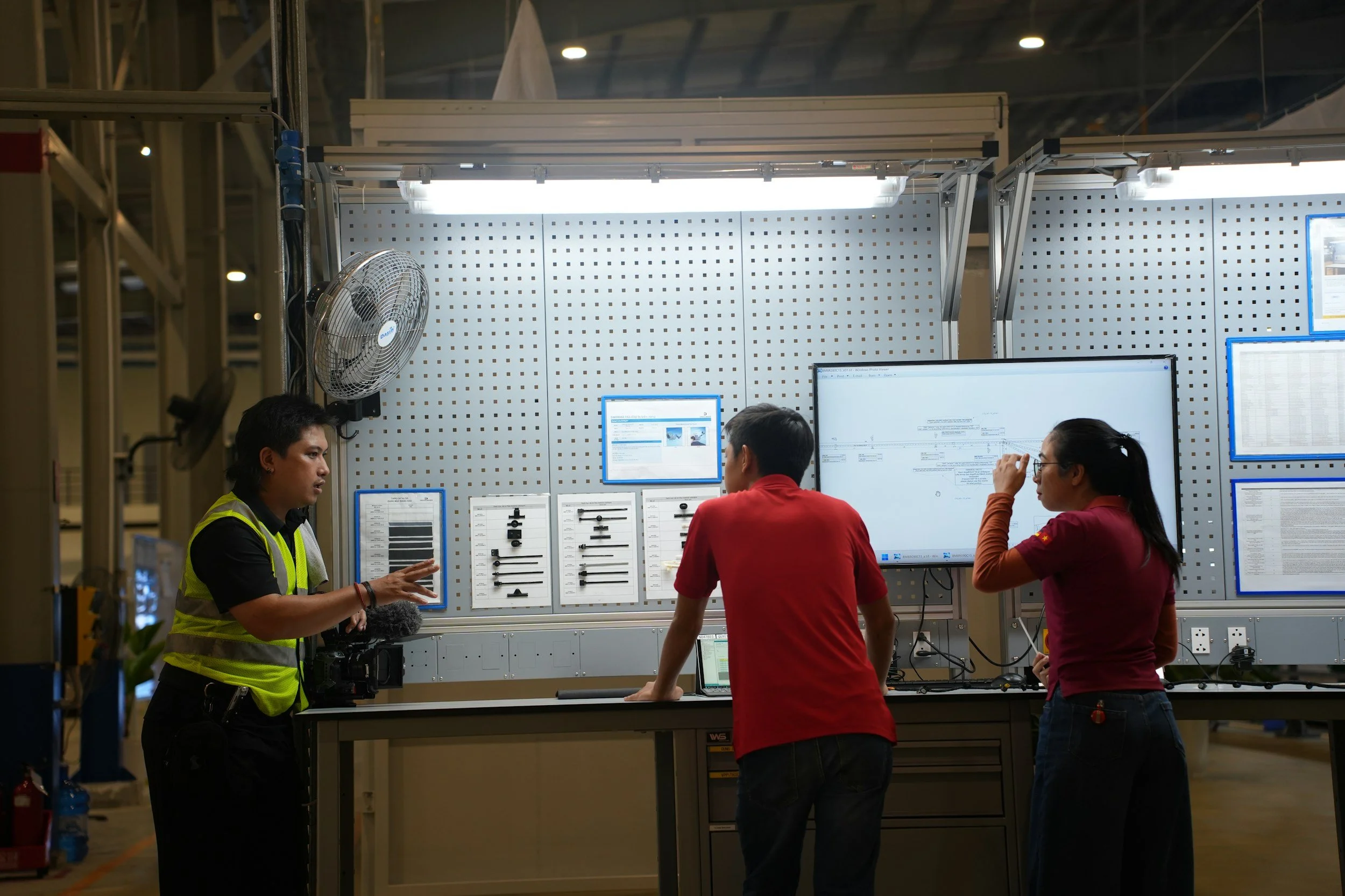 Three people in a workspace with large monitors and technical diagrams on a panel.