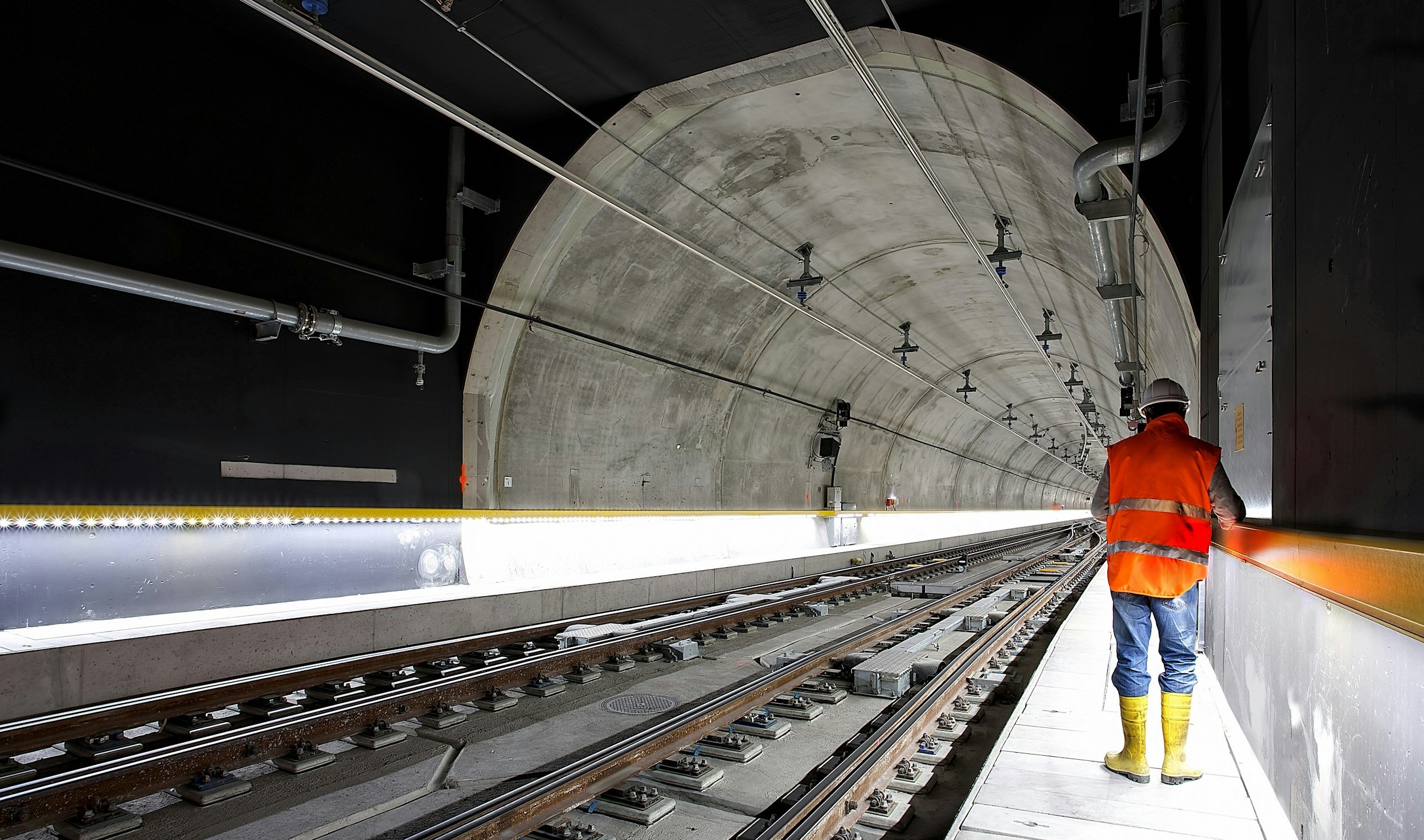 Construction worker in a bright orange safety vest, yellow rubber boots, and a white helmet walks along a tunnel with railway tracks, with equipment and piping overhead.