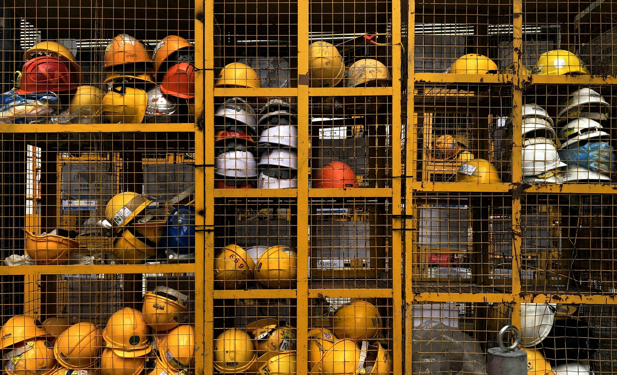 A large yellow metal storage cage filled with construction safety helmets of various colors including yellow, white, orange, red, and blue, some stacked and some hanging.