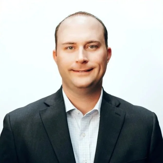 A portrait of a man with a receding hairline, wearing a dark suit and white collared shirt, against a plain white background.