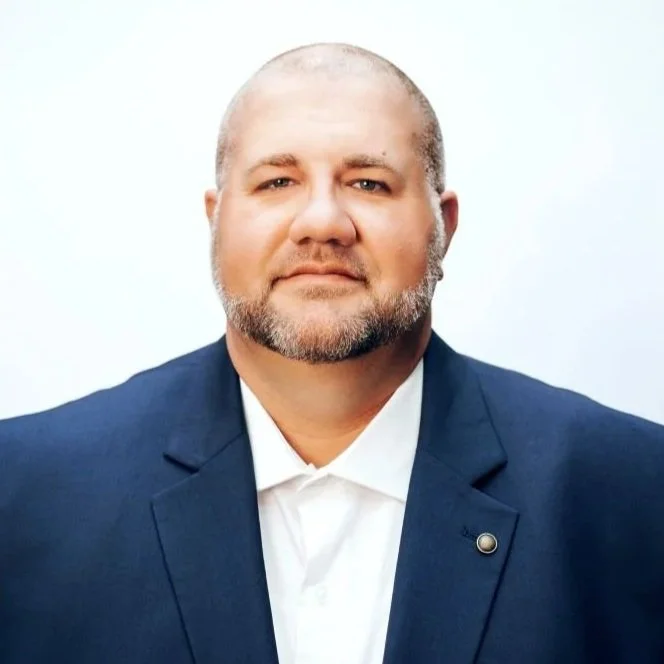 Headshot of a man with a beard and bald head, wearing a blue suit jacket and white shirt against a white background.