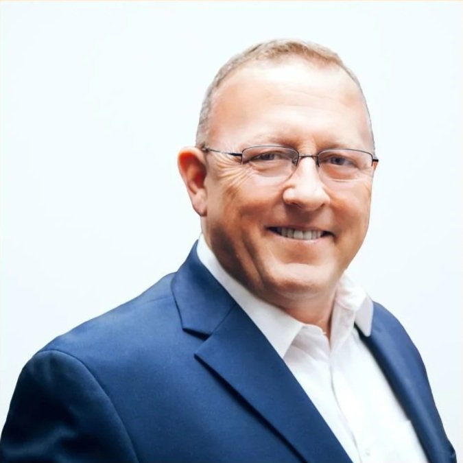 A smiling middle-aged man with glasses, wearing a white shirt and a blue suit jacket, standing against a plain light background.