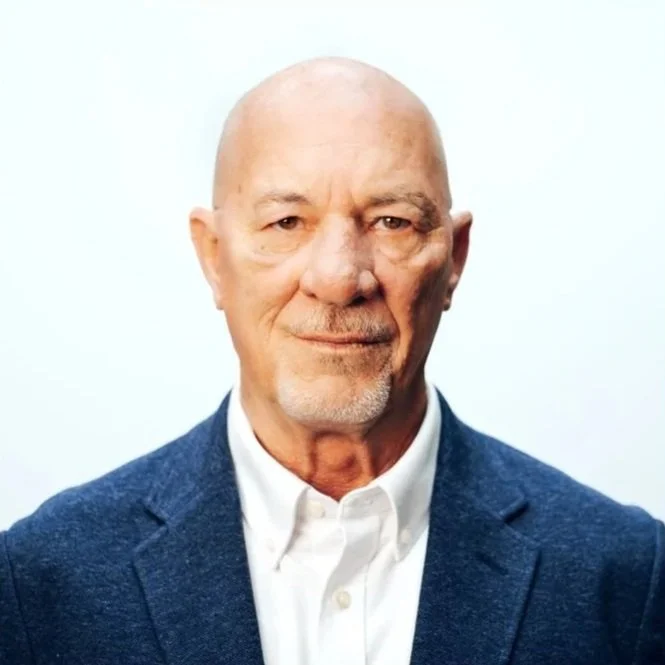 A portrait of an elderly man with a bald head, light facial hair, wearing a blue blazer and white shirt, against a plain white background.