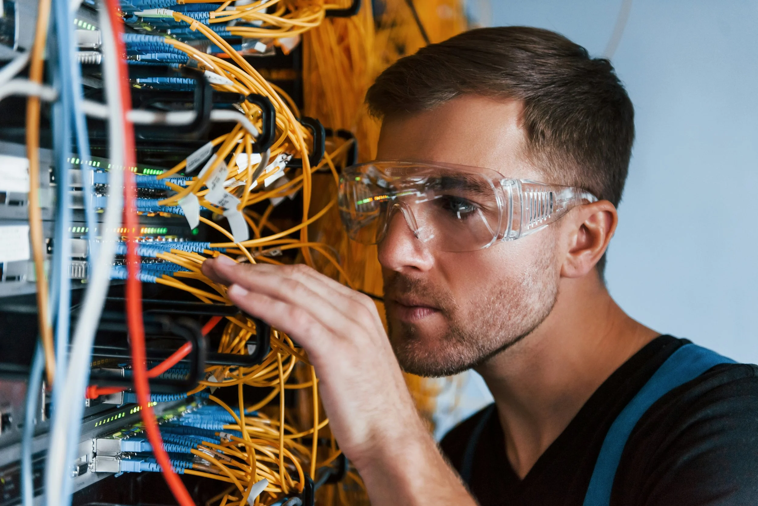 A man wearing safety glasses working with networking equipment and yellow cables in a server room.