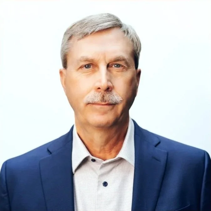 Portrait of an older man with gray hair and a mustache, wearing a blue suit jacket and a white shirt, standing against a plain white background.