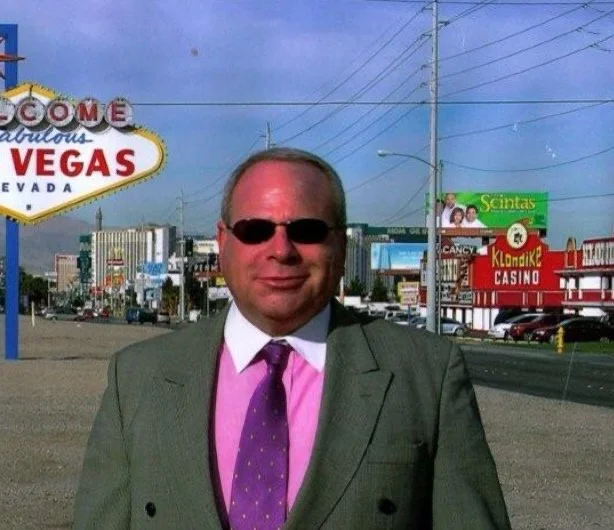 A man in a gray suit, pink shirt, and purple tie standing in front of the Las Vegas Strip with signs for casinos and the 'Welcome to Fabulous Las Vegas' sign visible in the background.