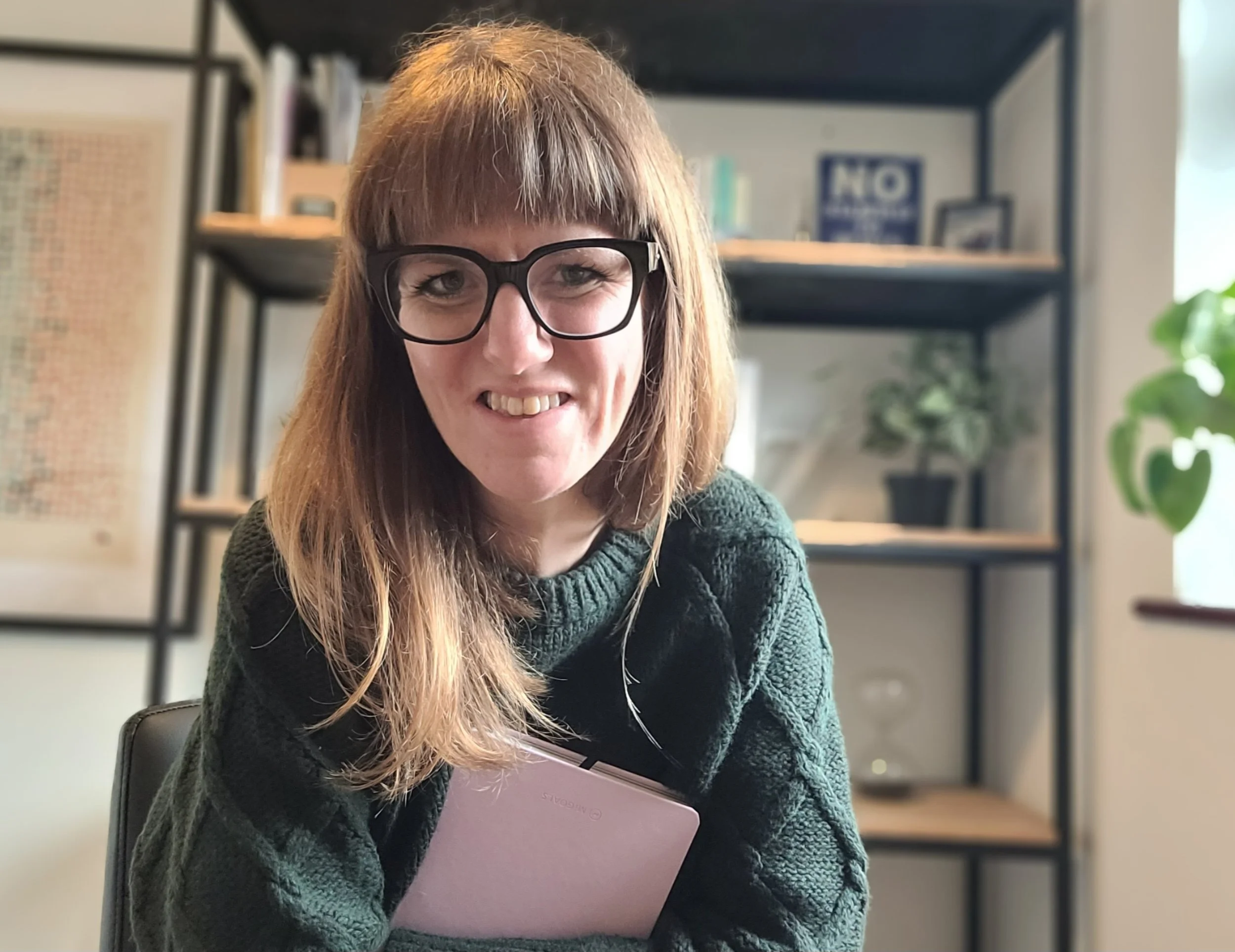 A picture of Charlotte Buxton. She has long light brown hair, glasses, and a green jumper. She is smiling while holding a pink diary in a cozy office with a bookshelf, potted plant, and framed artwork in the background.