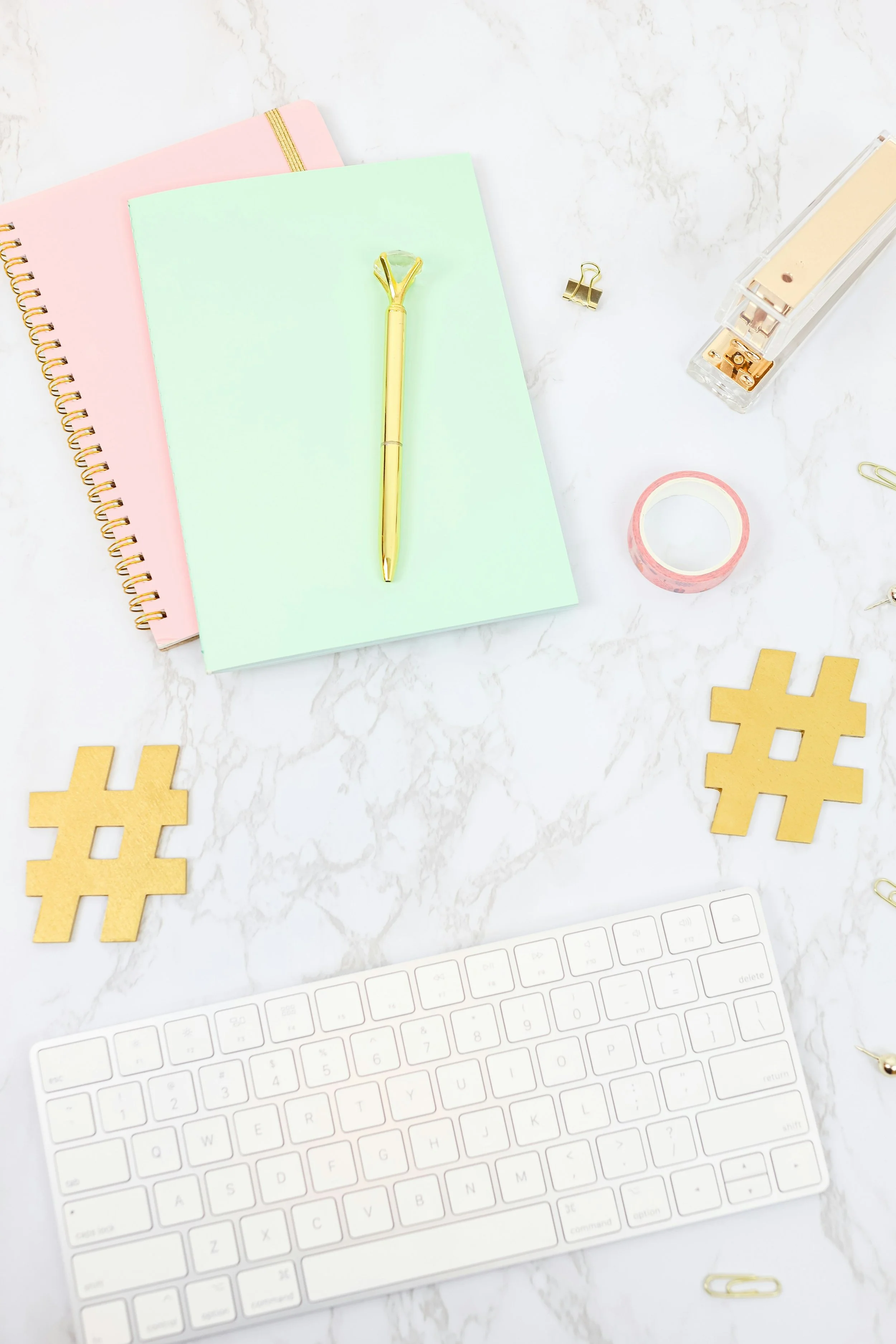 A flat lay of a workspace on a white marble surface, including pastel notebooks, a gold pen, a roll of pink tape, gold hashtag symbols, a white wireless keyboard, and various gold paper clips and a stapler.