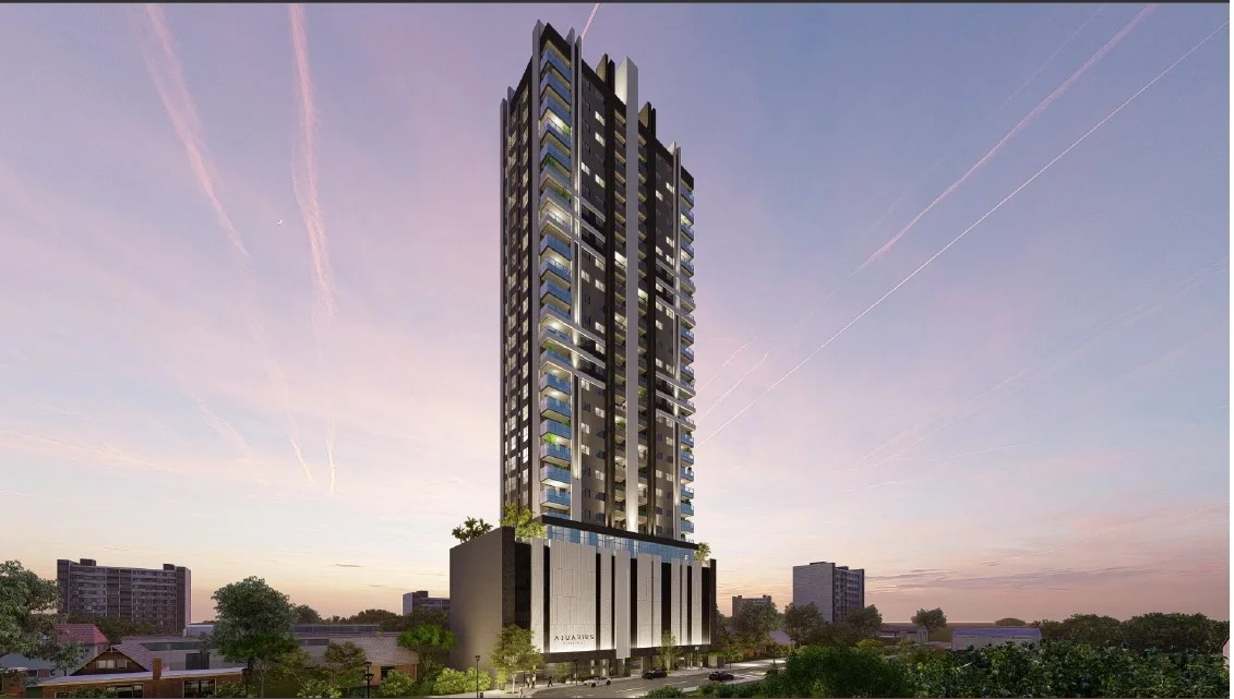 Prédio moderno de alta torre com iluminação noturna, árvores ao redor e pôr do sol ao fundo.